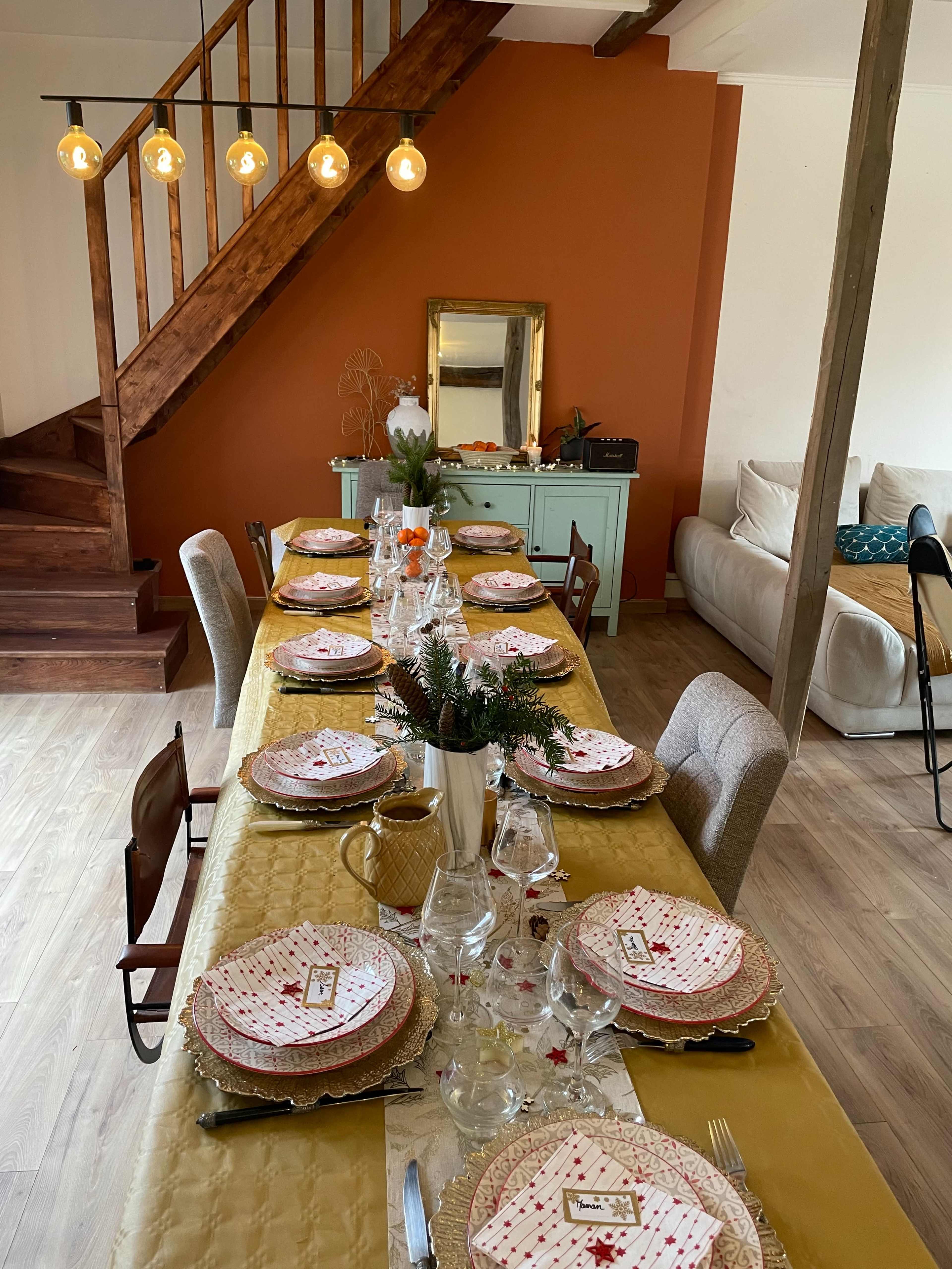 A dining table is set with patterned plates and glassware in a warmly lit room featuring a staircase and an orange accent wall.