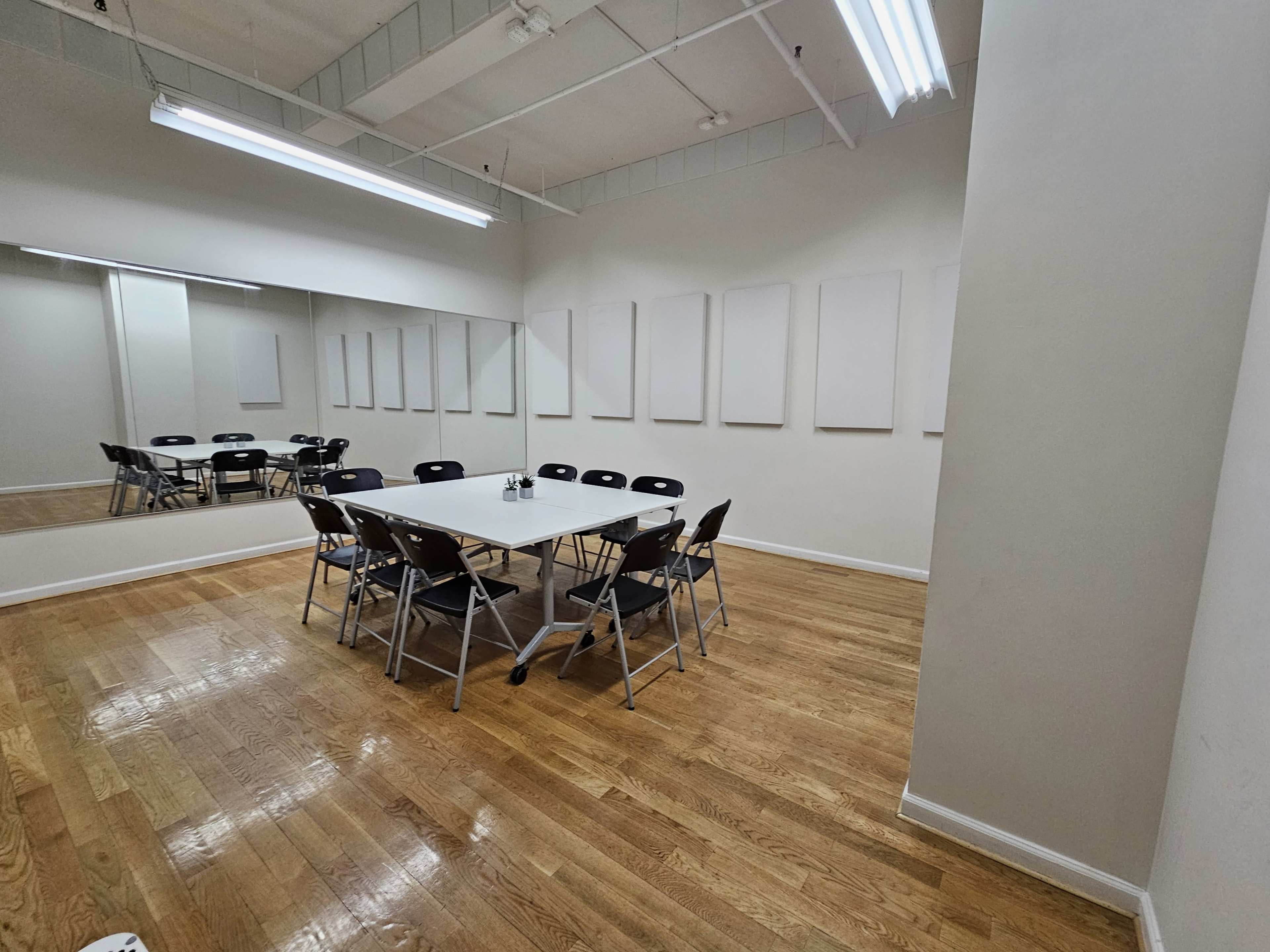 A meeting room with a rectangular table surrounded by folding chairs, illuminated by fluorescent lights, and featuring empty white walls.