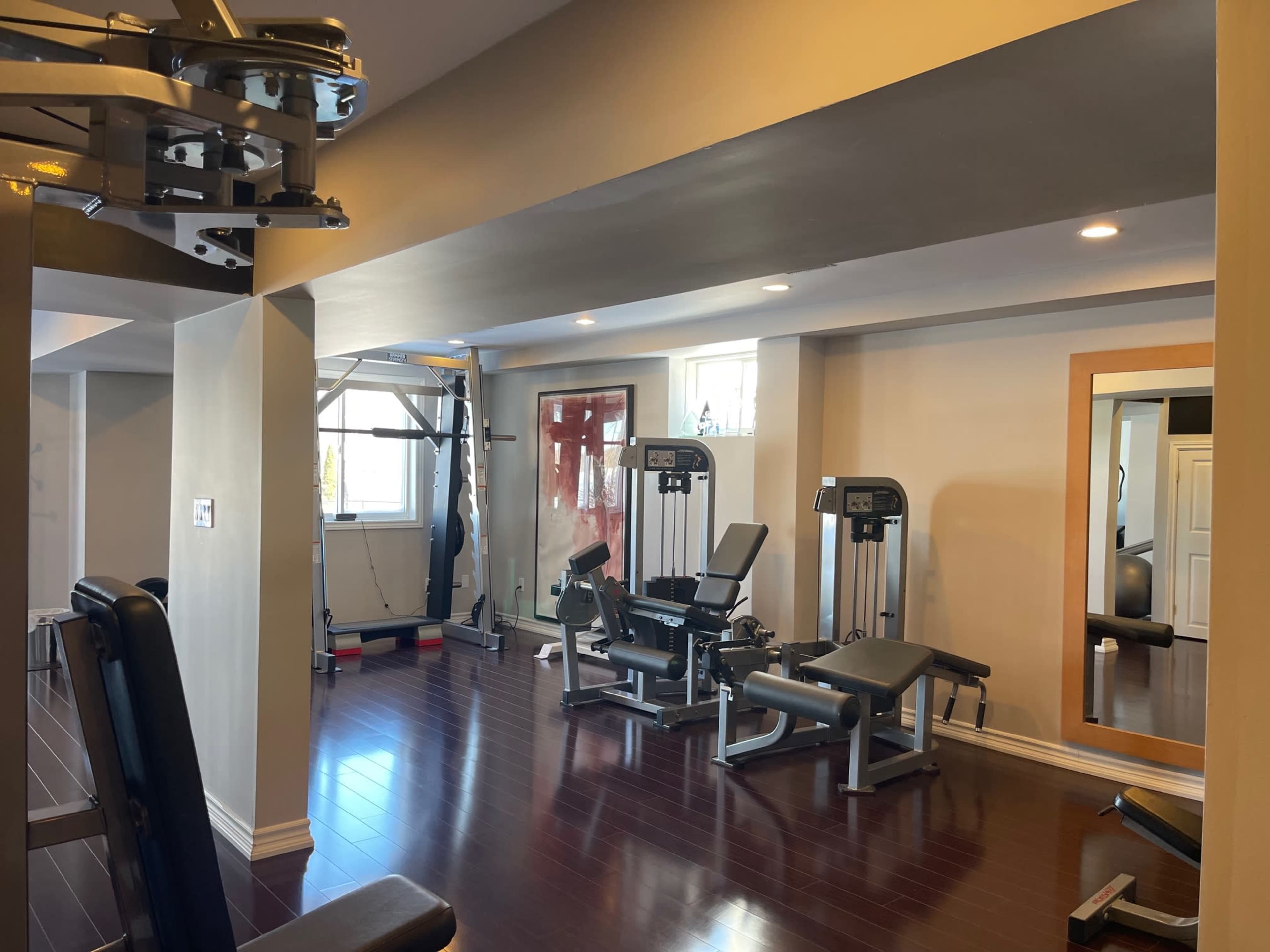 The image shows a well-lit home gym featuring various exercise equipment, including weight machines and benches, with wooden flooring and large windows.