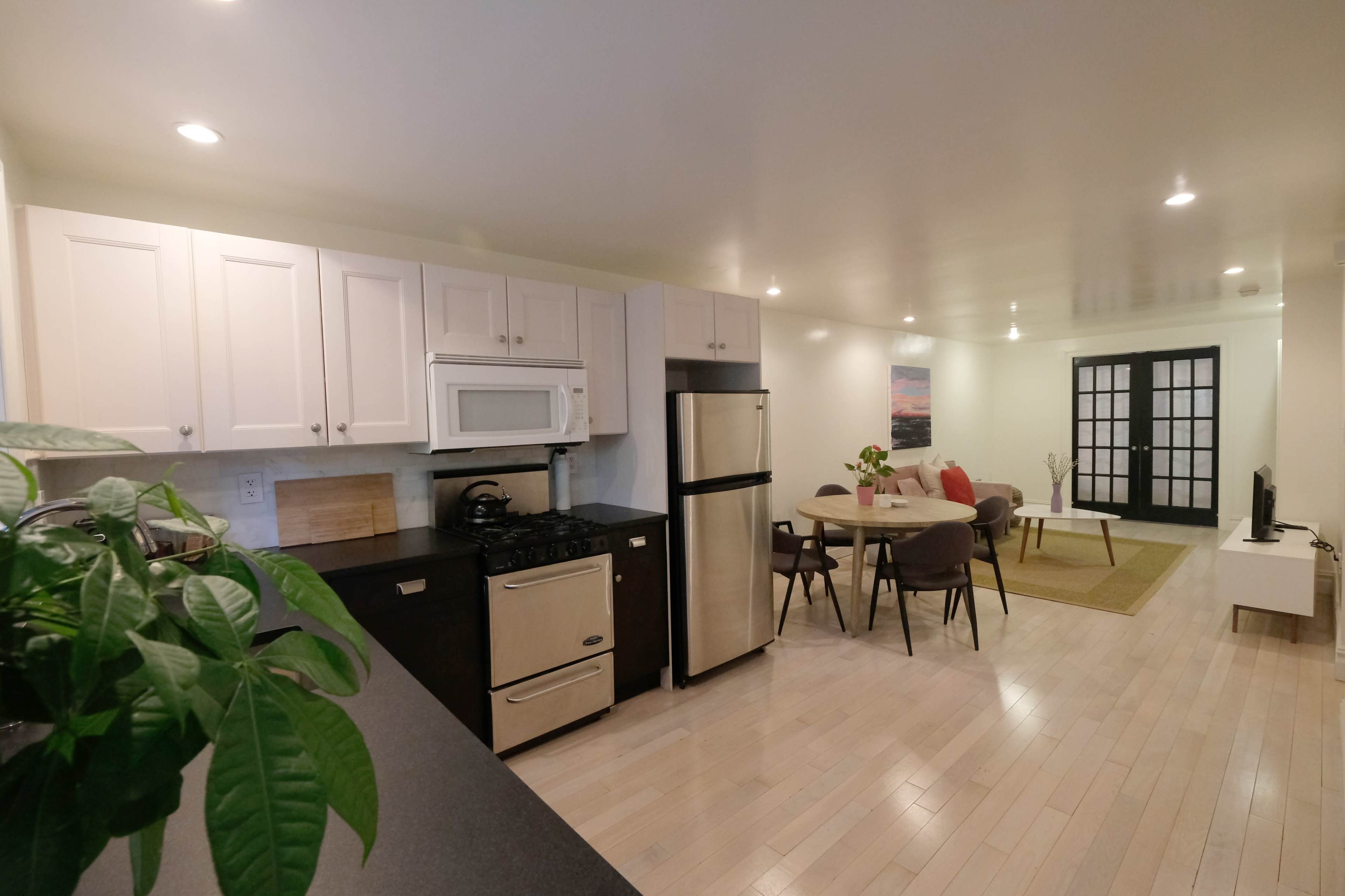 The image shows a modern kitchen and living area with white cabinetry, stainless steel appliances, a circular dining table, and a seating arrangement.