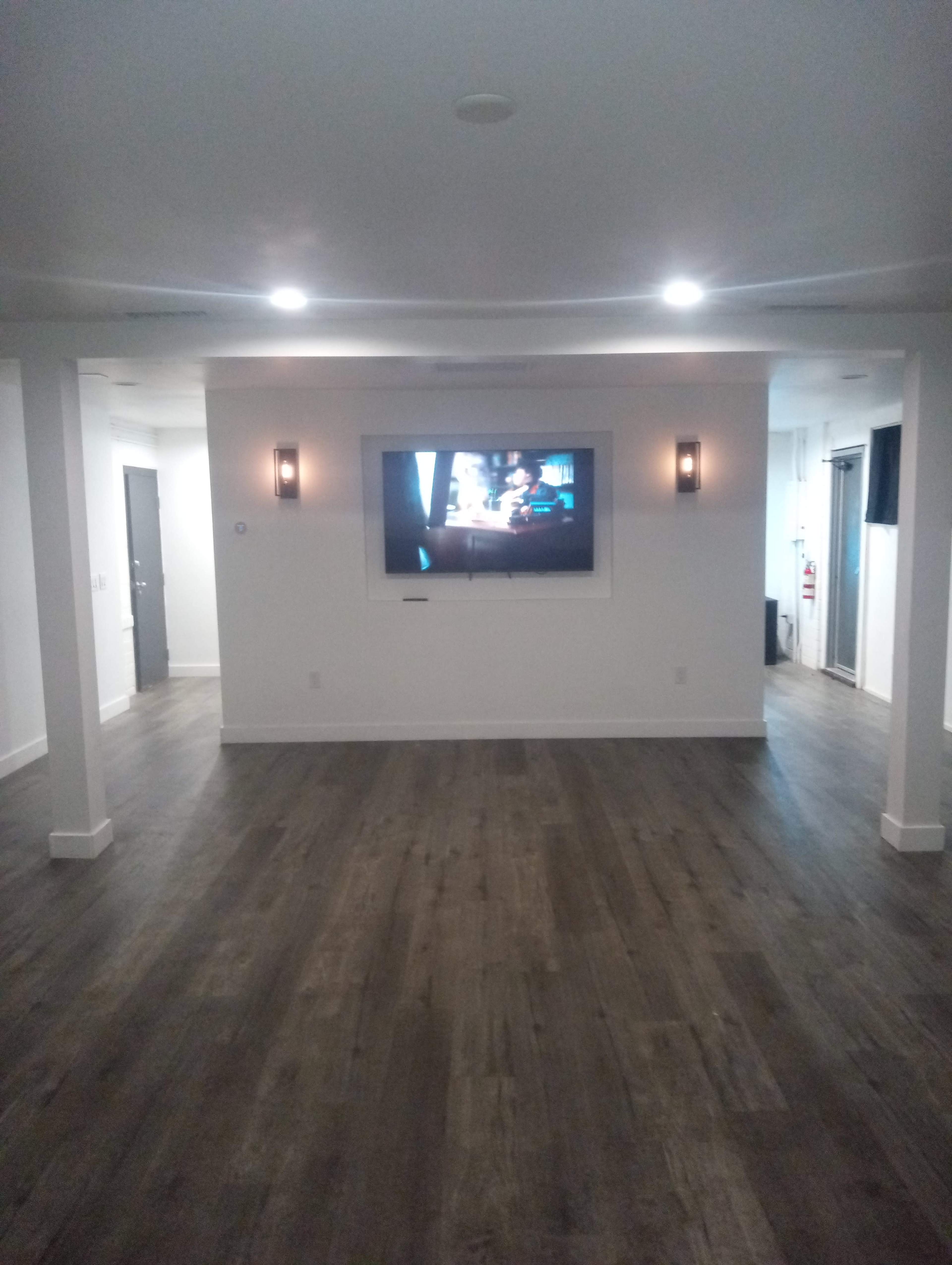 The image shows a spacious, well-lit room with a large flat-screen TV mounted on the wall and wooden flooring.