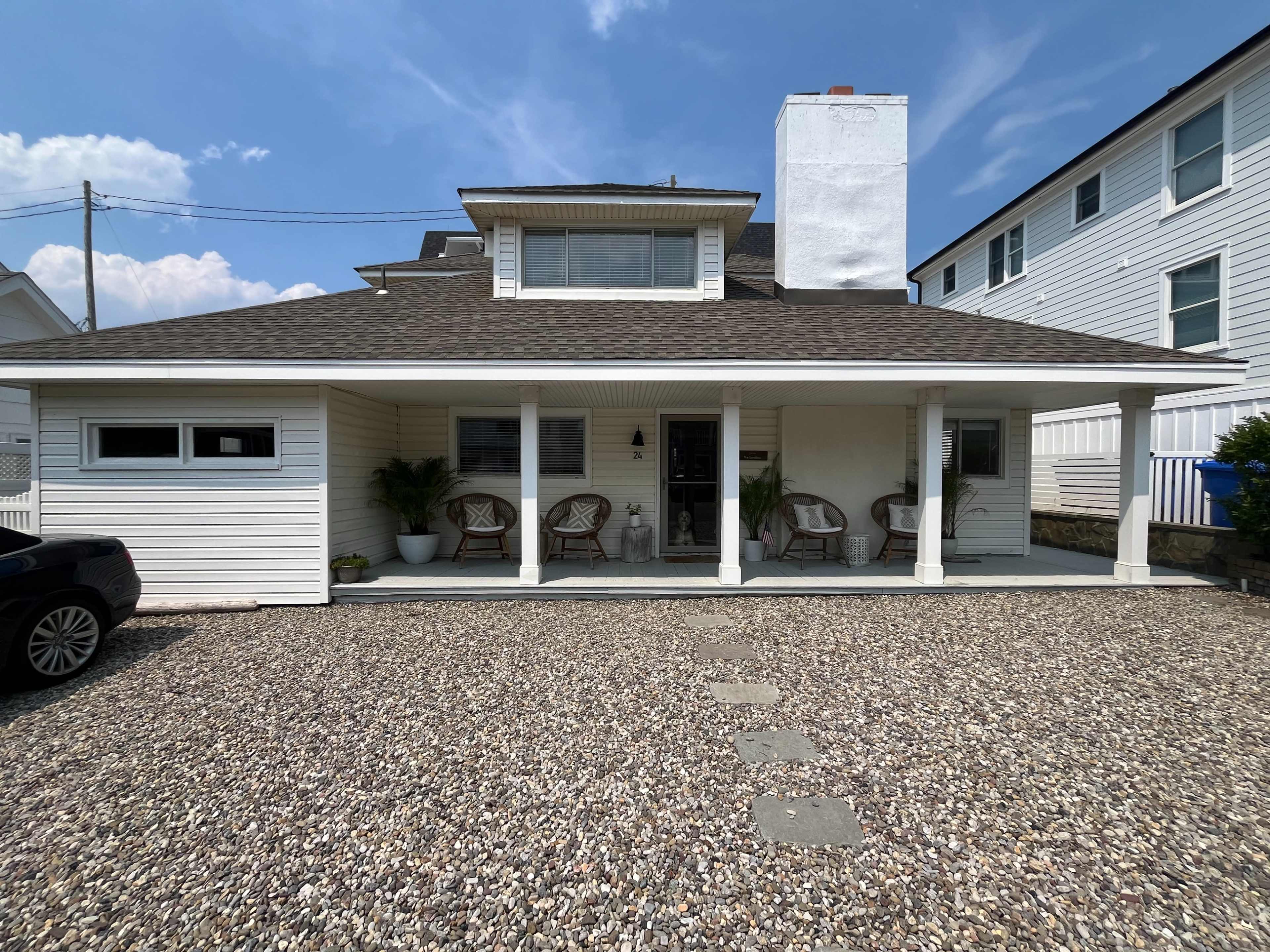A two-story house with a front porch supported by white columns, featuring a chimney and a gravel driveway lined with decorative stones.