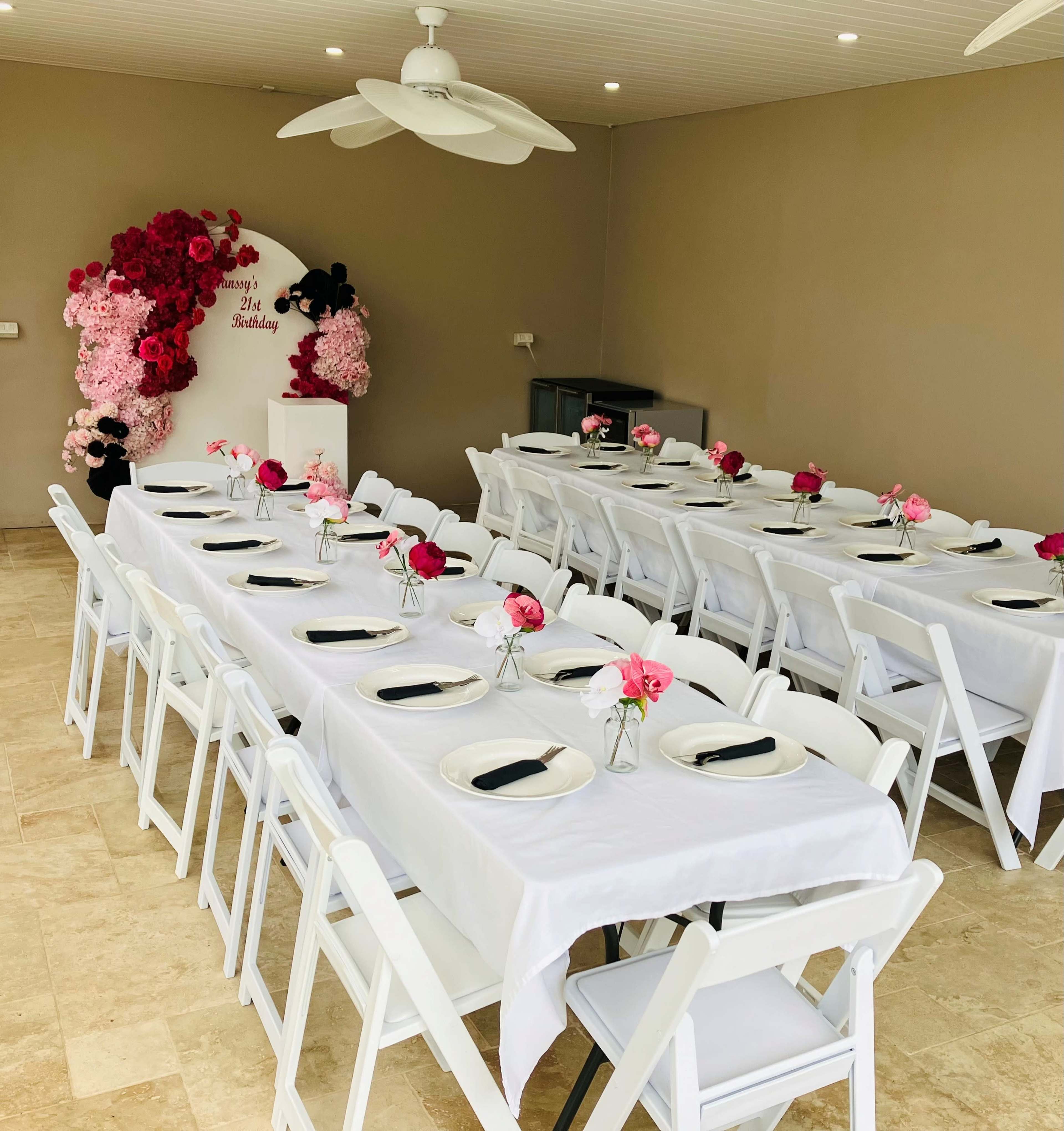 A spacious dining area is set with long tables covered in white tablecloths, each adorned with floral centerpieces and black napkins, beneath a decorative wall of pink flowers.
