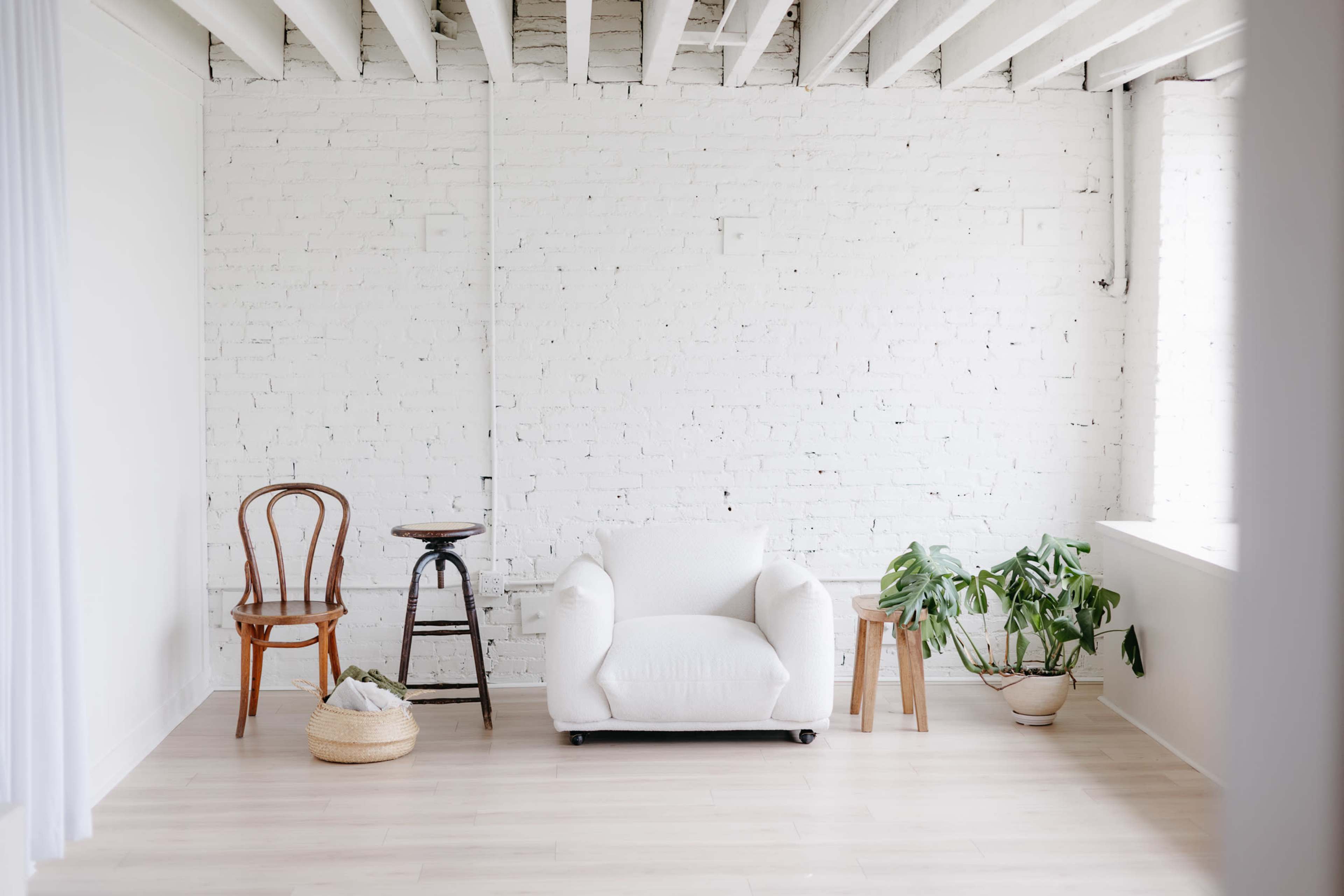 The image features a minimalist interior with a white brick wall, a white armchair, a wooden chair, a small table, and several indoor plants.
