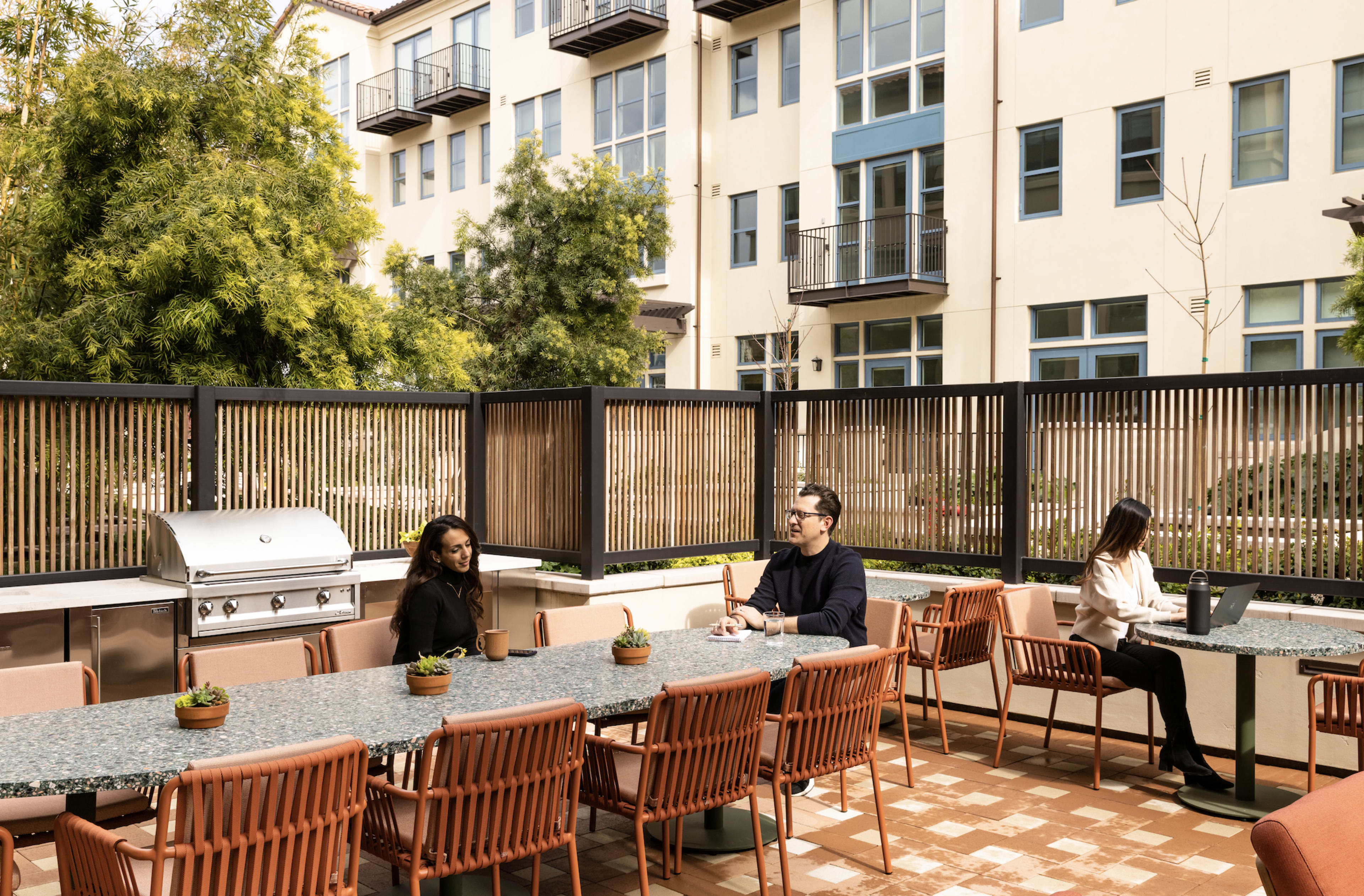 A group of three people sits at a long outdoor dining table in a shared courtyard with a barbecue grill and surrounding greenery.