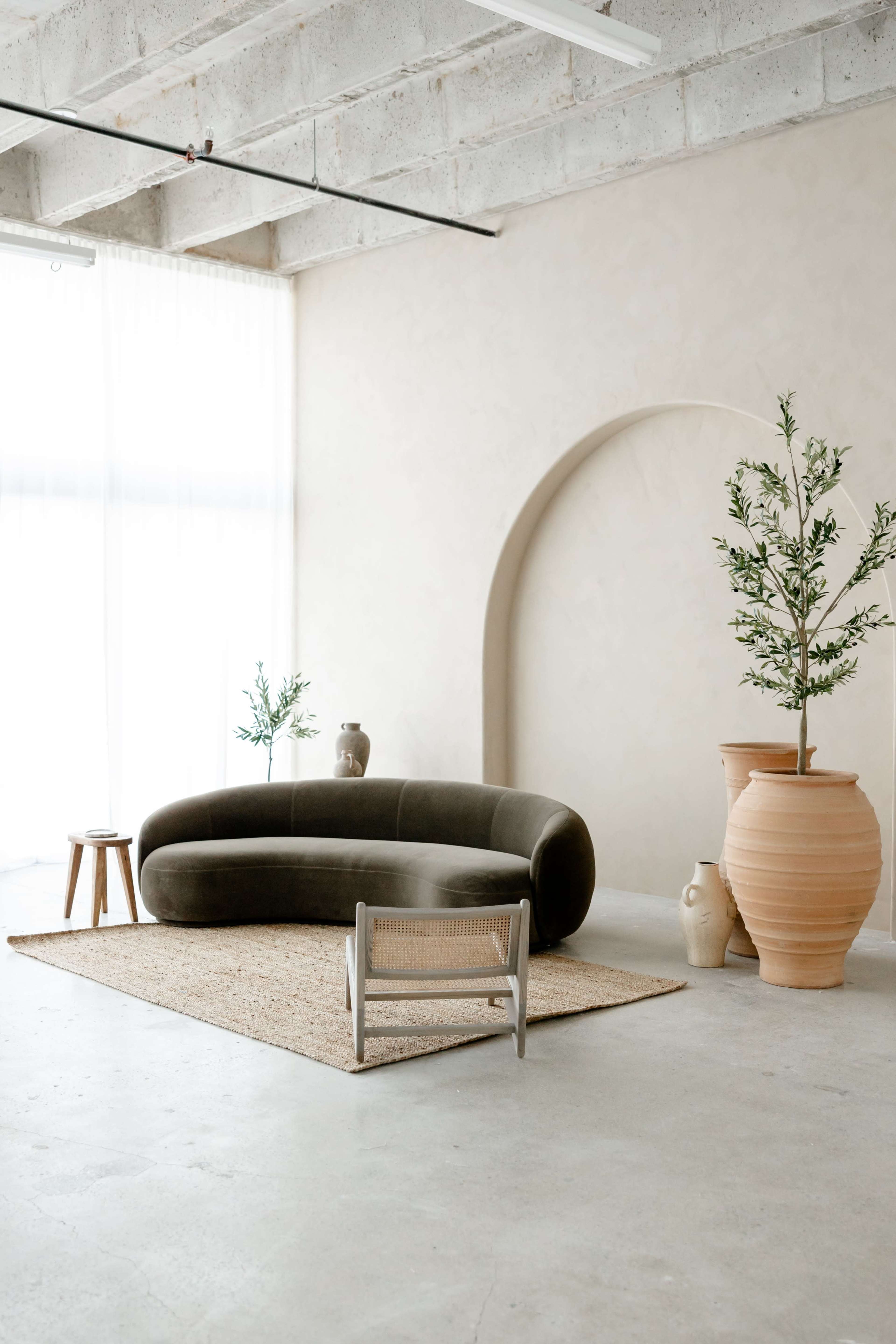 The image shows a modern interior with a curved green sofa, a small wooden table, and large ceramic pots, all set against a light-colored wall and natural light filtering through sheer curtains.
