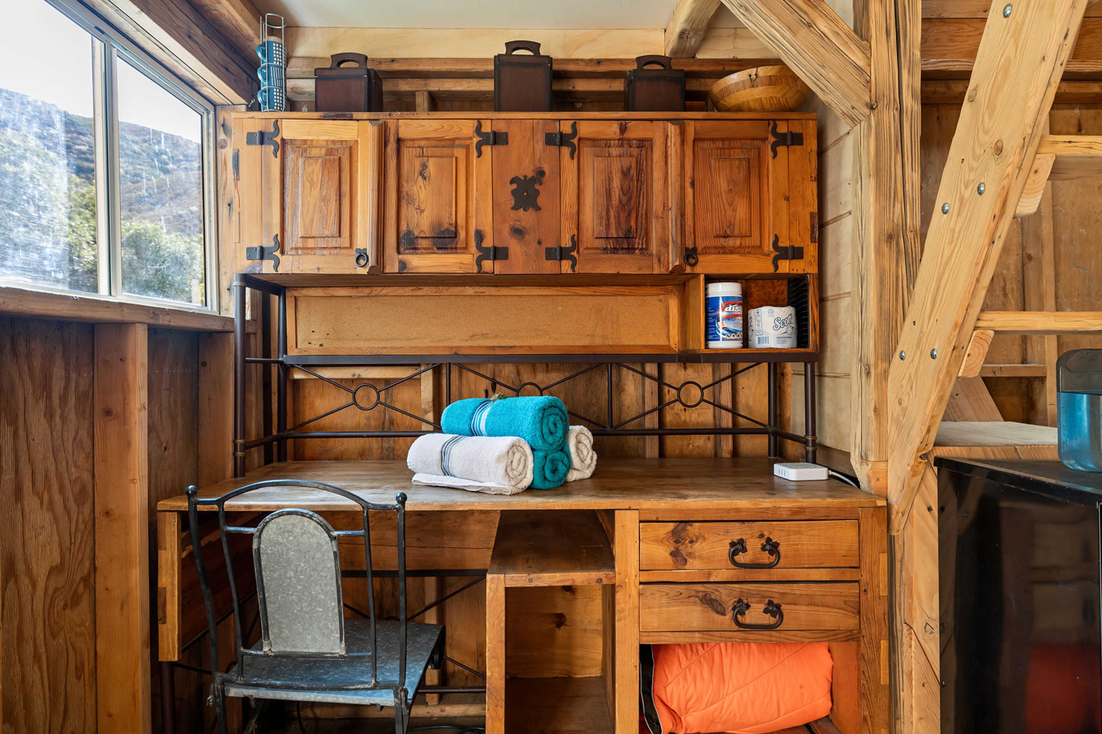 The image shows a wooden desk with a chair, neatly arranged towels, and cabinets set in a rustic interior space with natural light coming through a window.