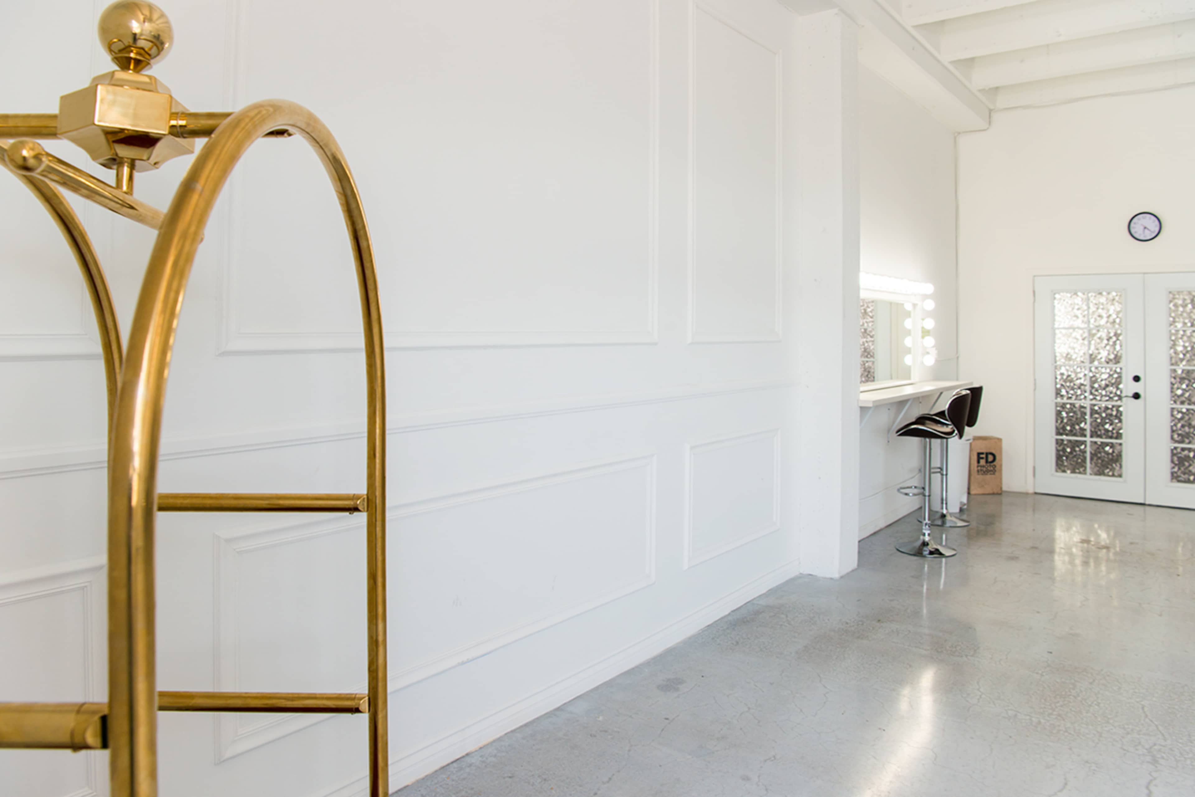 A bright, minimalist room with white walls, a bar table with chairs, a large mirror with lights, and a brass clothing rack.