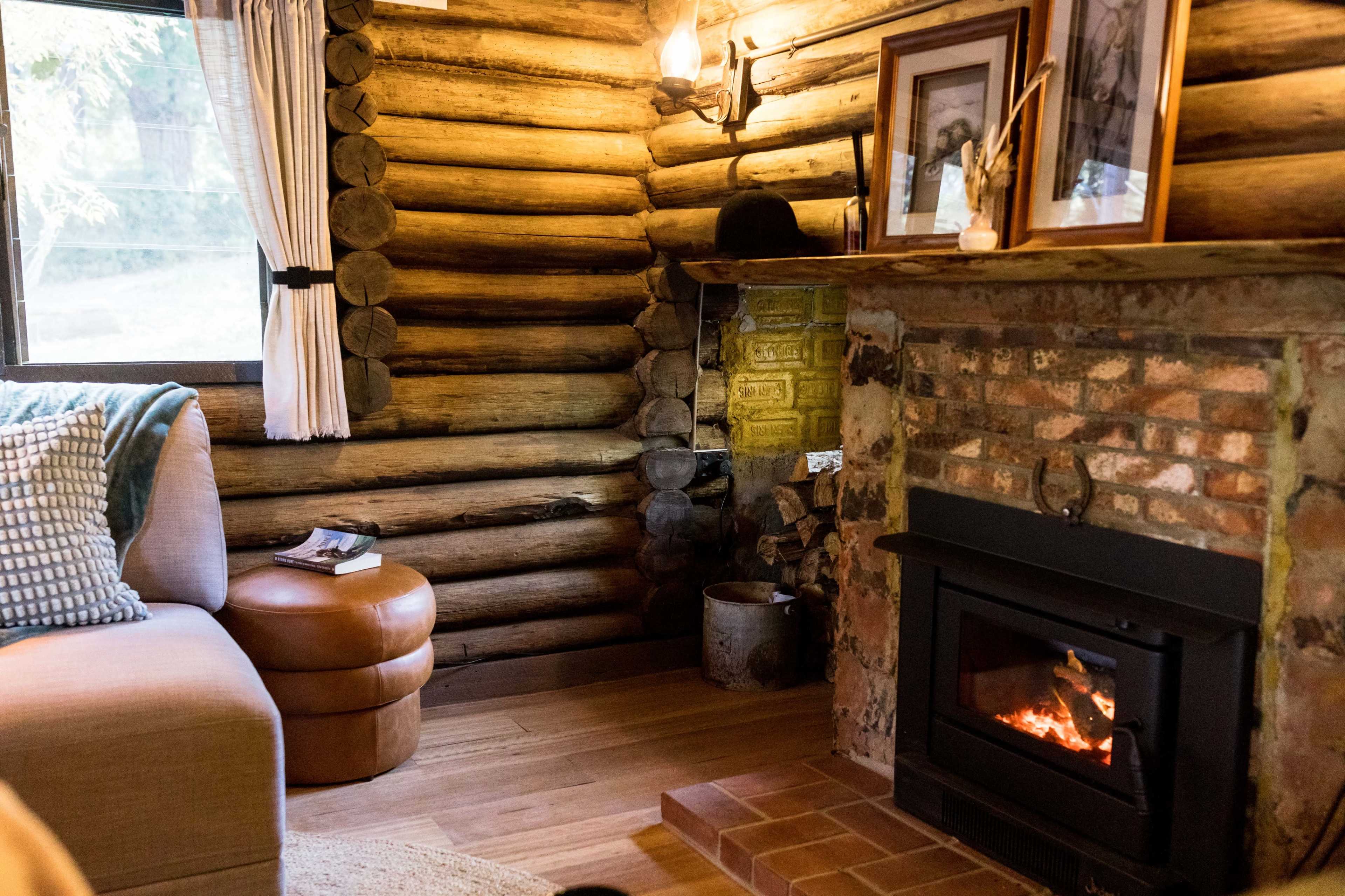 A cozy living room with a wooden log wall, a brick fireplace with a small fire, a round leather ottoman, and framed pictures on the mantel.