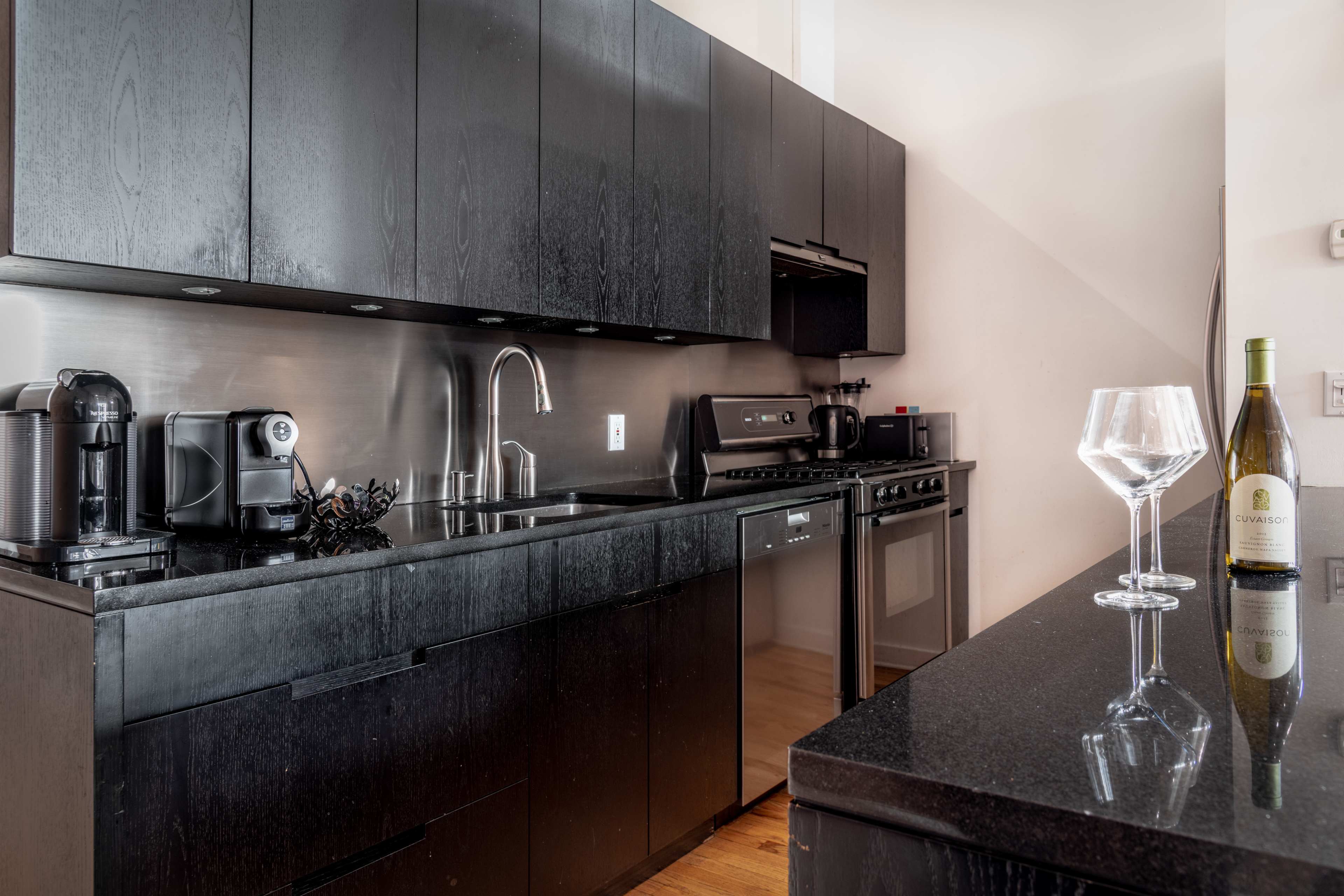 A modern kitchen with dark cabinetry, stainless steel appliances, and a black countertop featuring two wine glasses and a bottle of wine.