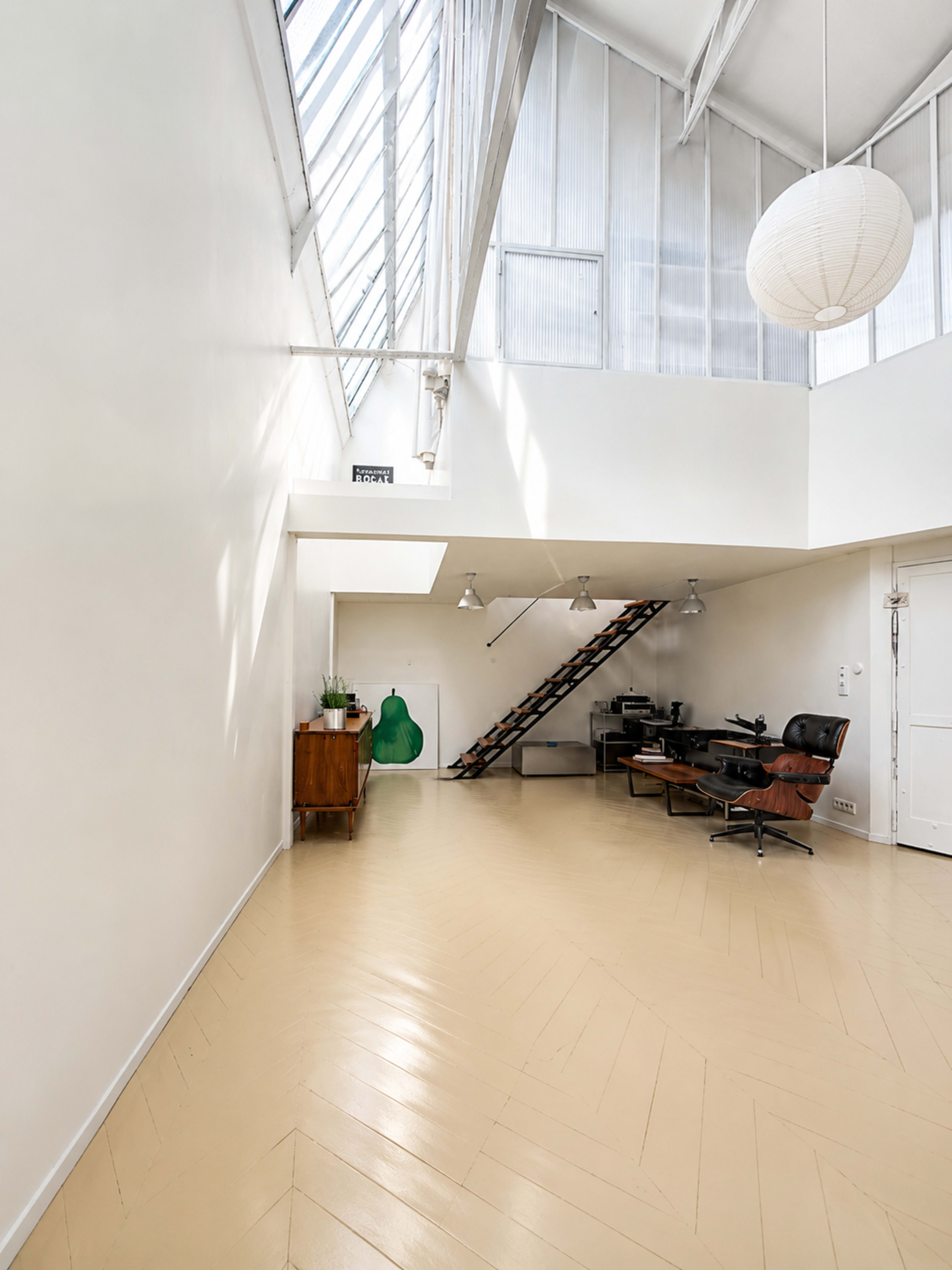 The image shows a spacious, minimalist interior with high ceilings, featuring a staircase, a workplace setup, and a green artwork on the wall.