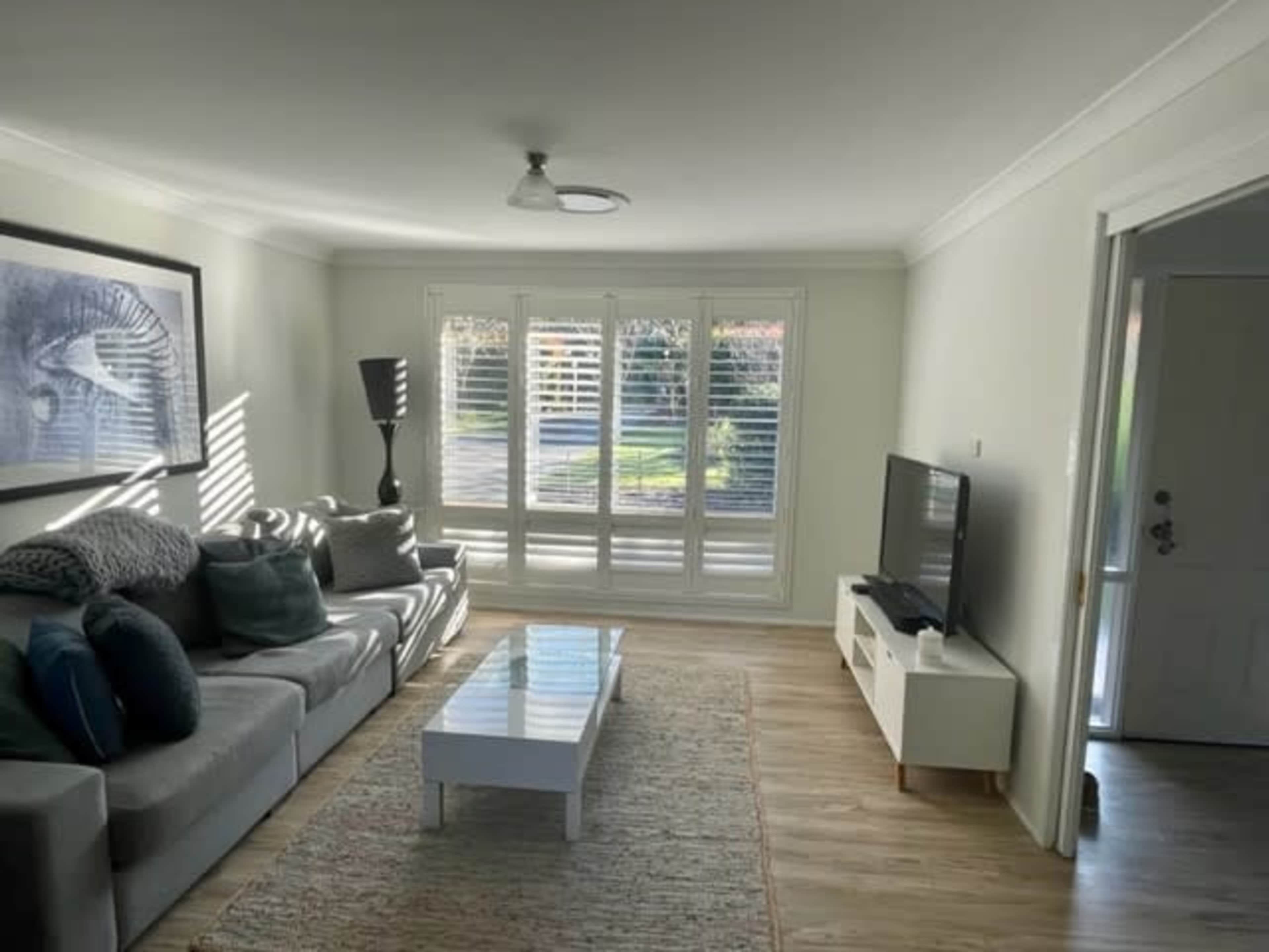 The living room features a gray sectional sofa, a white coffee table, a TV on a media console, and large windows with plantation shutters allowing natural light.