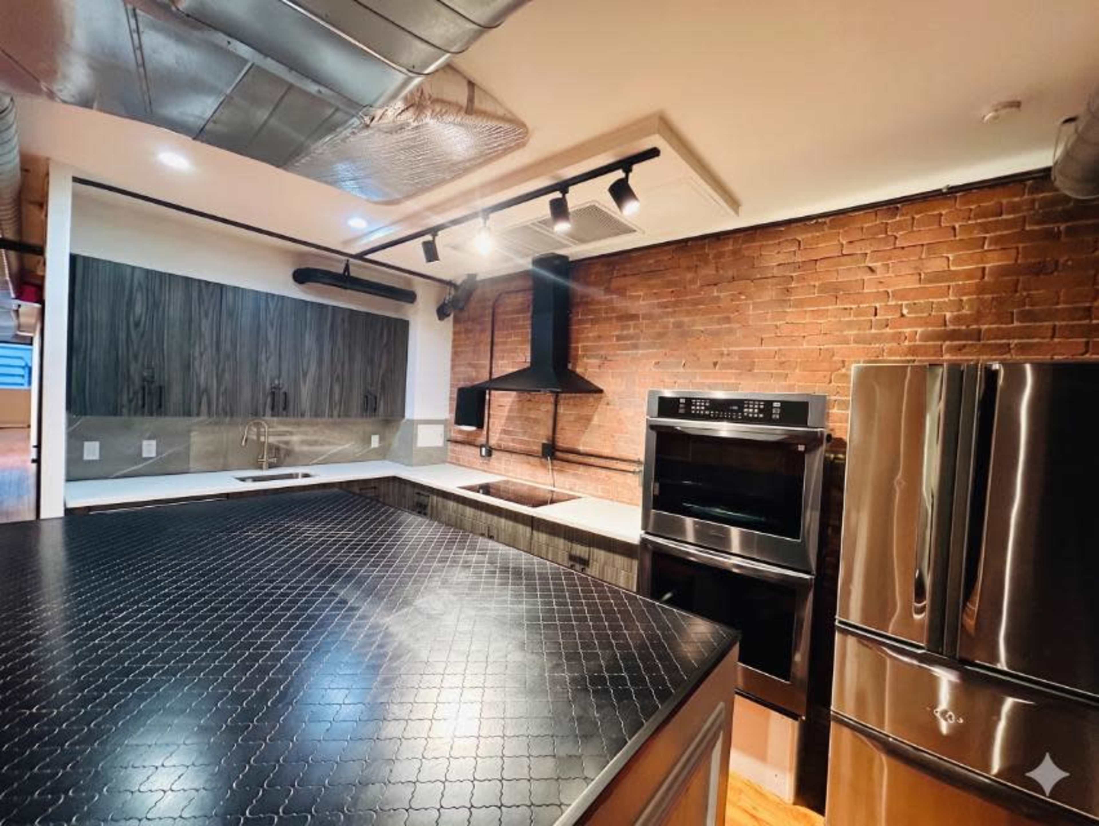The image shows a modern kitchen featuring a dark countertop, stainless steel appliances, and exposed brick walls.