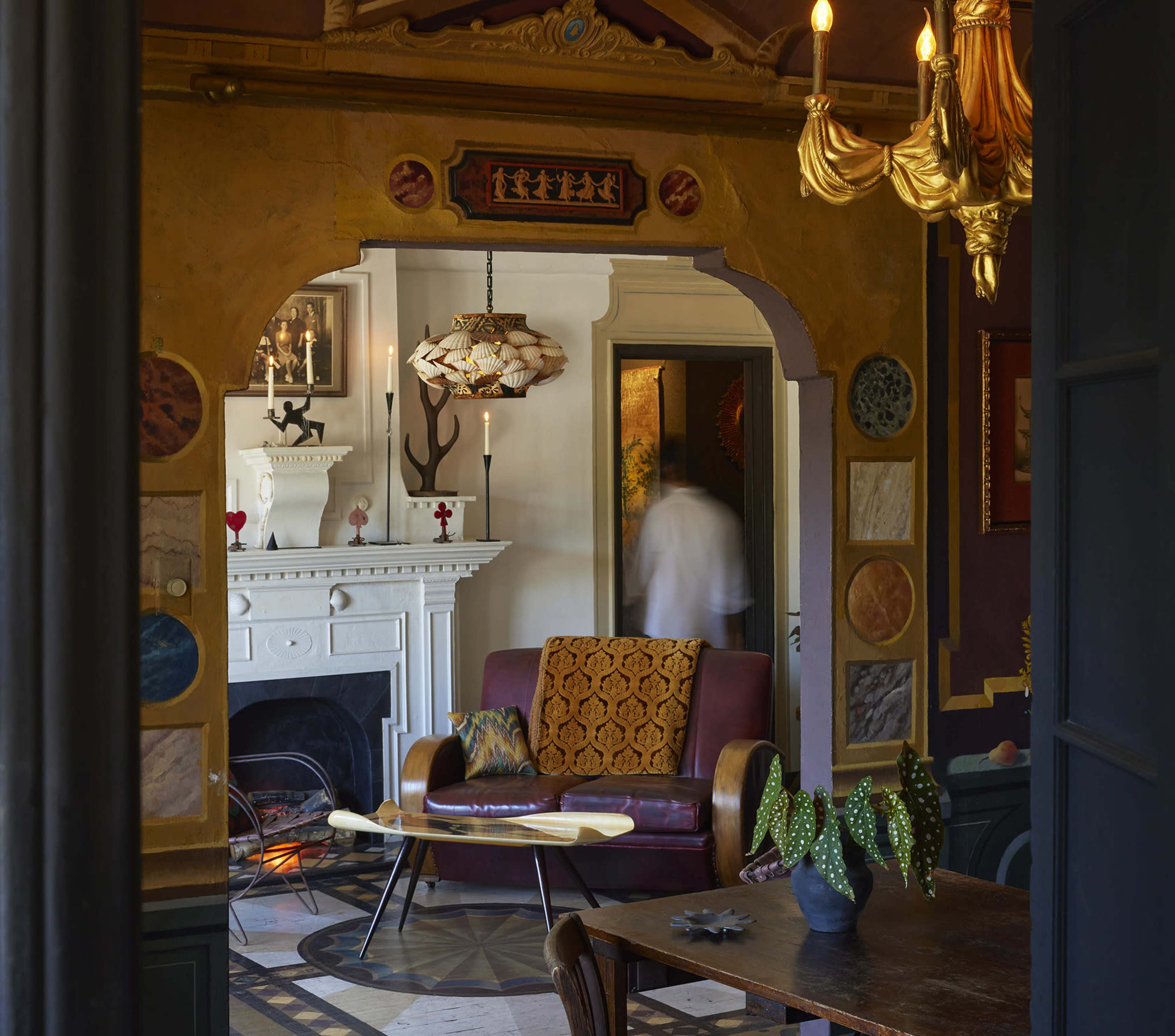 The image shows a living room with a maroon sofa, patterned cushions, an ornate archway, and a fireplace, framed by decorative wall elements and a table with a cactus centerpiece.