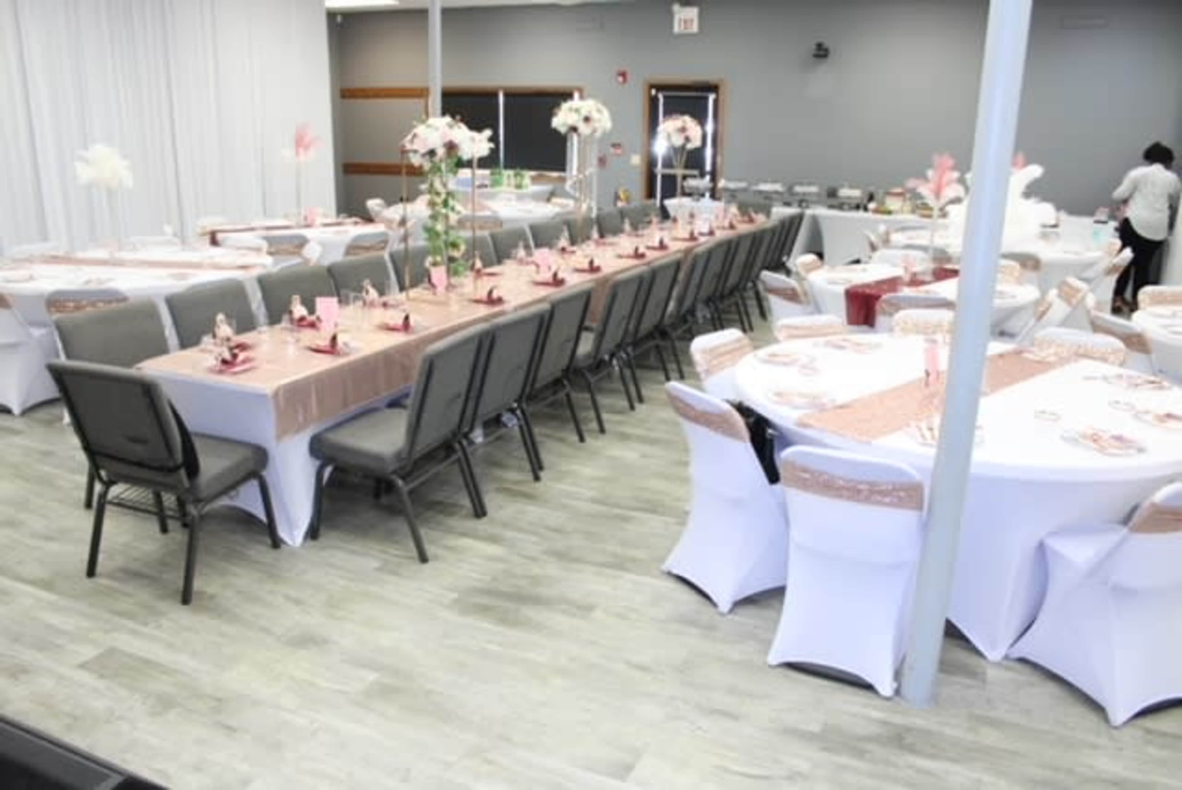 A banquet hall arranged with multiple tables covered in white and beige tablecloths, decorated with flower centerpieces and pink accents.