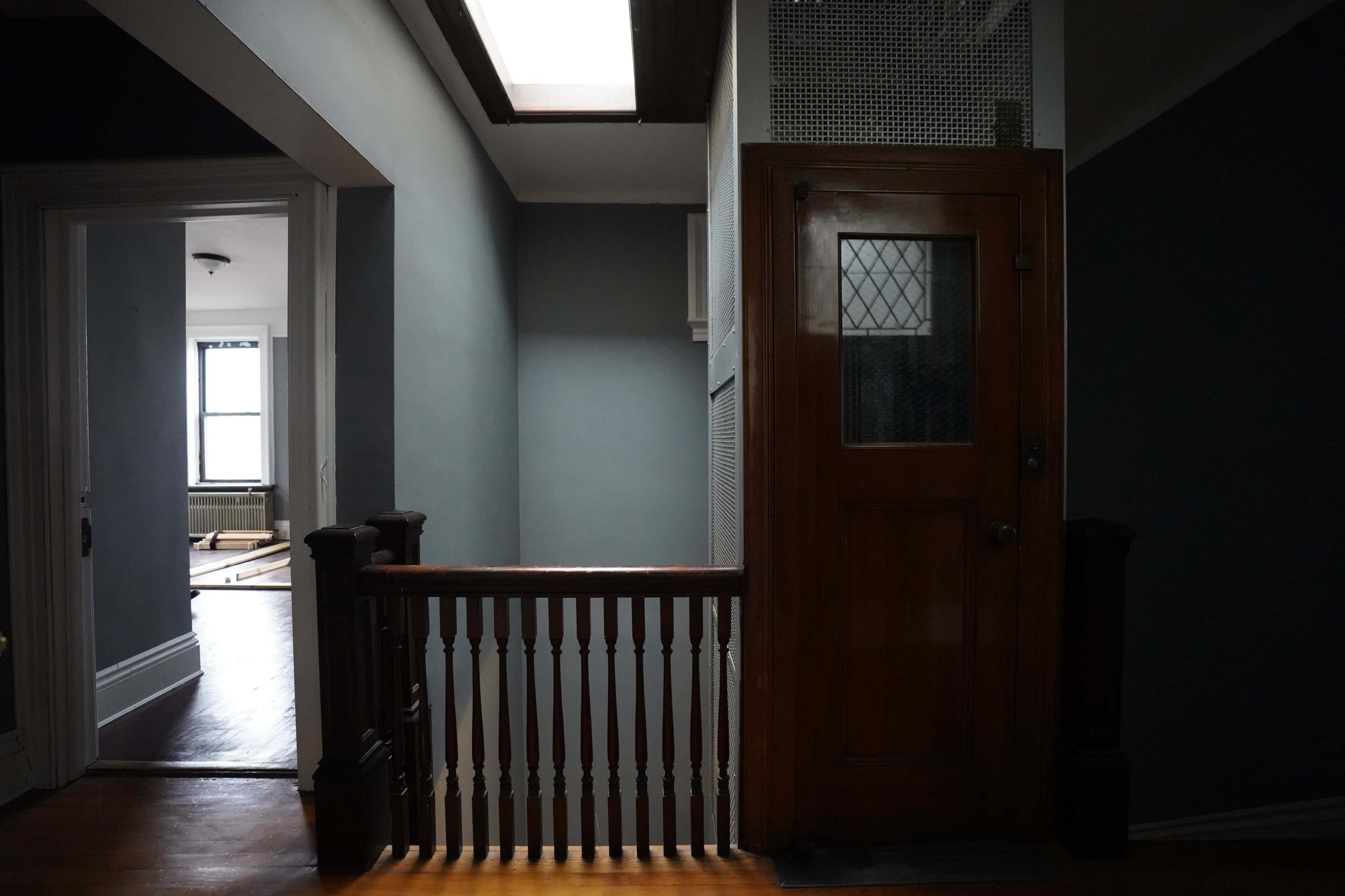 A dimly lit hallway with a wooden railing leading to a closed door, and an open doorway on the left that opens to a brighter room.