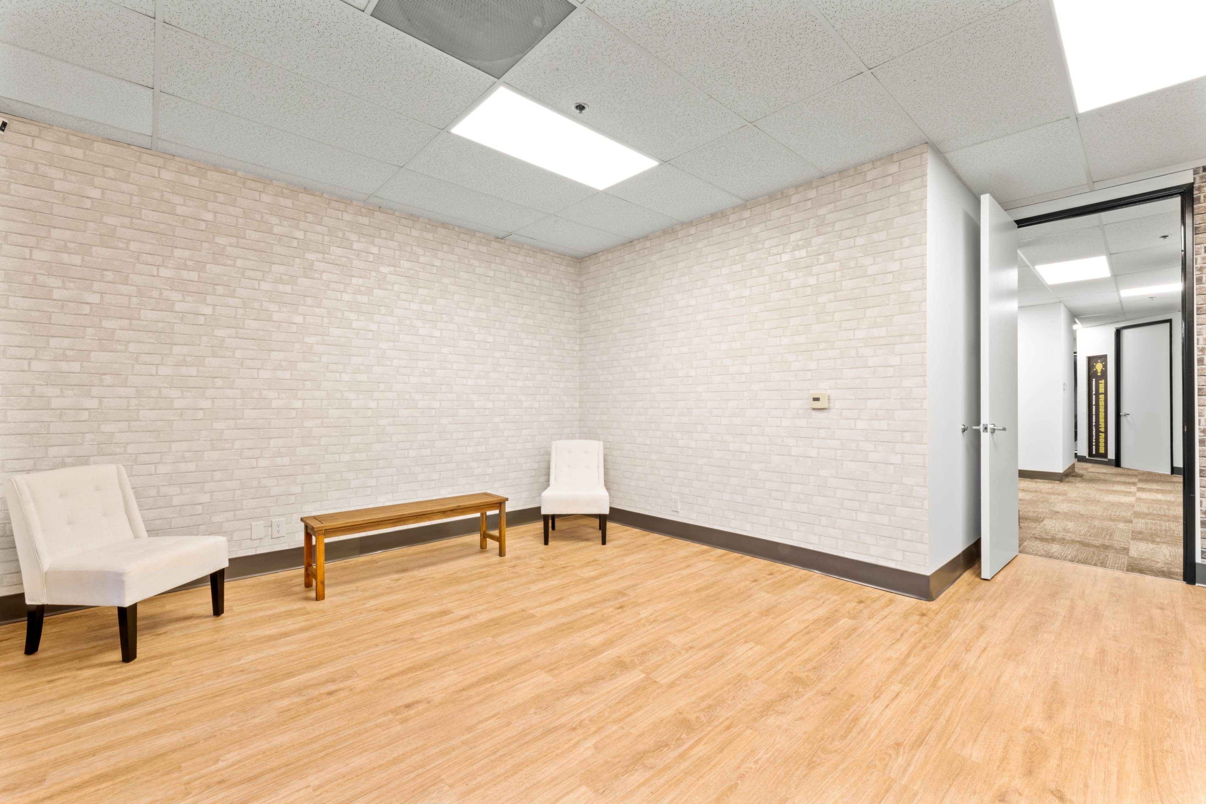 The image shows a sparsely furnished room with two white chairs and a wooden coffee table, featuring light-colored brick walls and a door leading to another area.