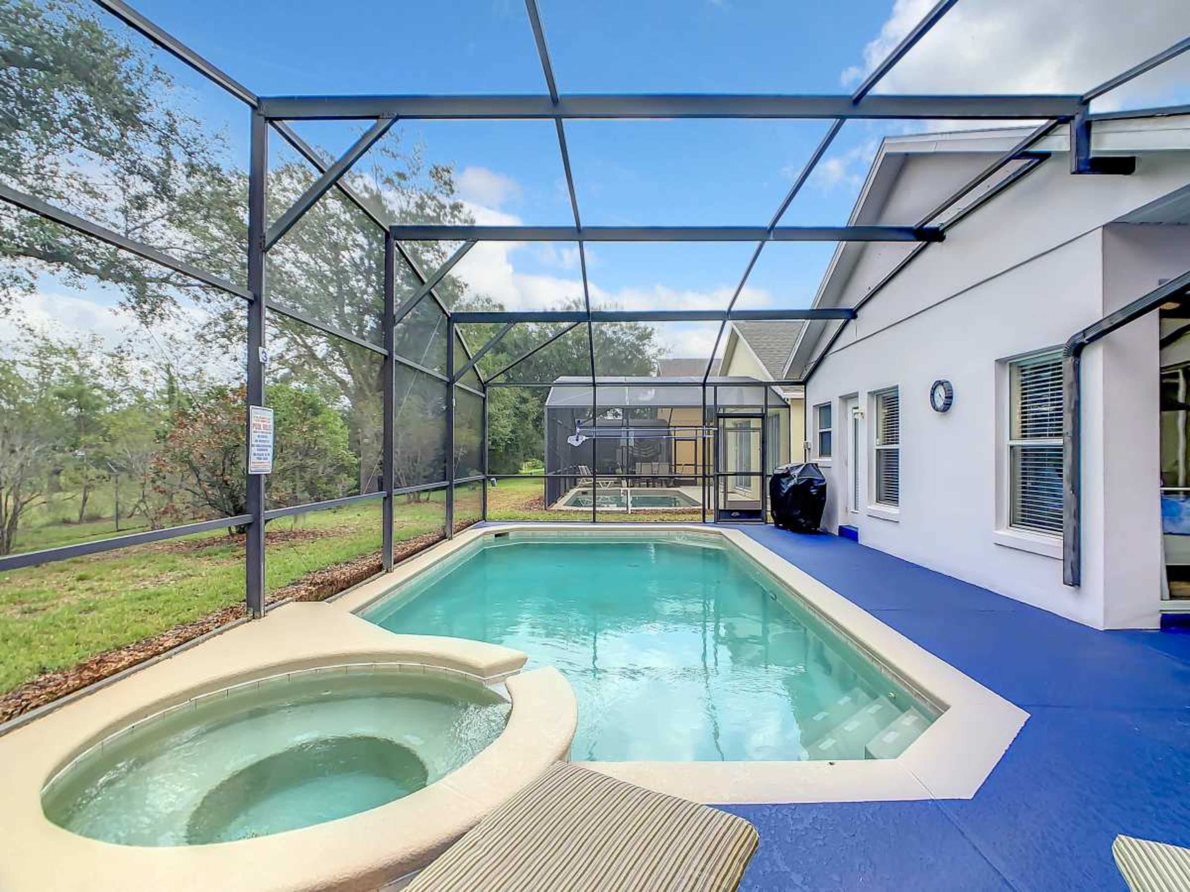 The image shows a backyard swimming pool with a spa, surrounded by a screened enclosure, and a view of a grassy area beyond.