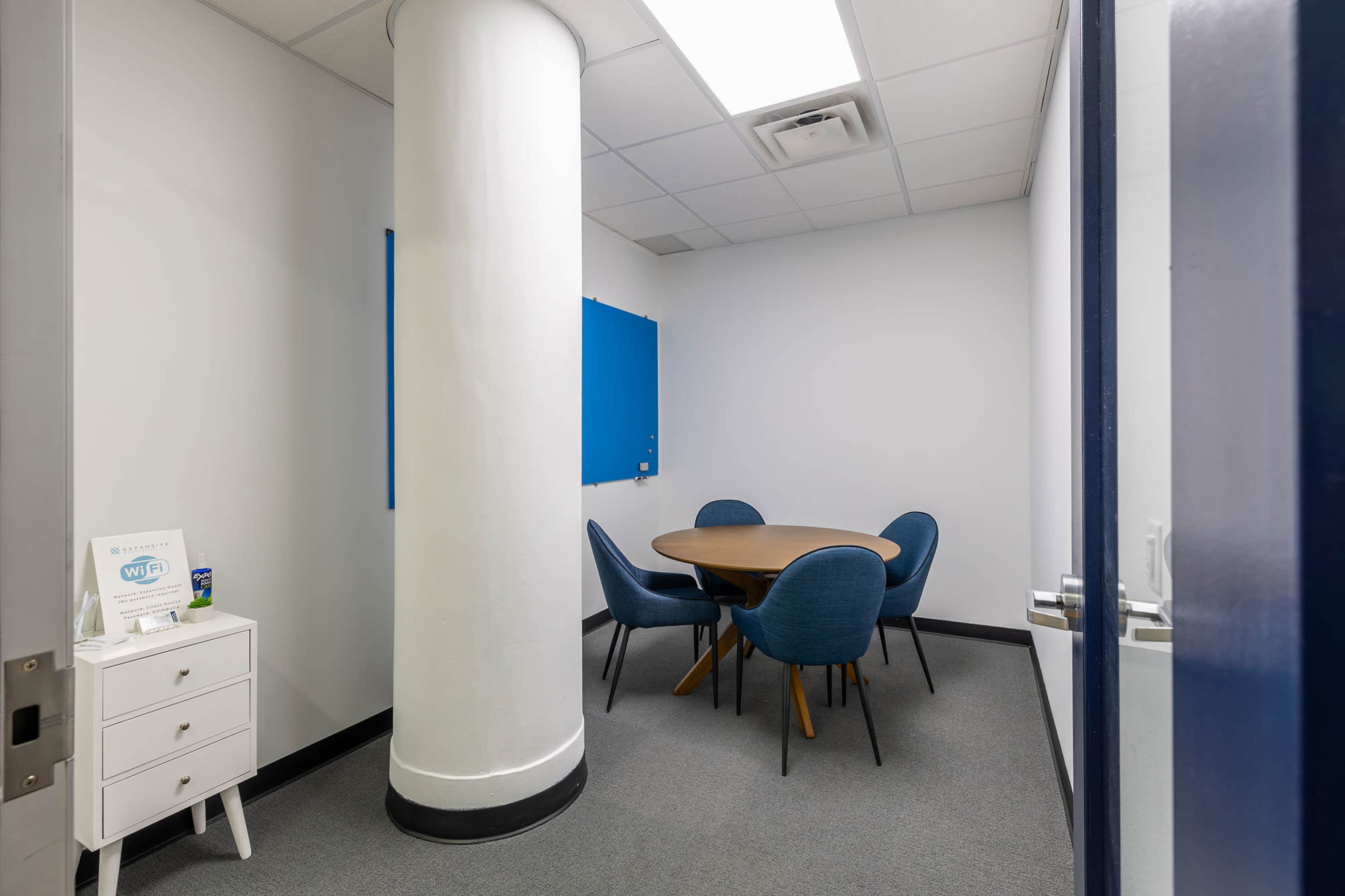 The image shows a small conference room featuring a round wooden table surrounded by four blue chairs, with a whiteboard and a Wi-Fi sign on a side table.