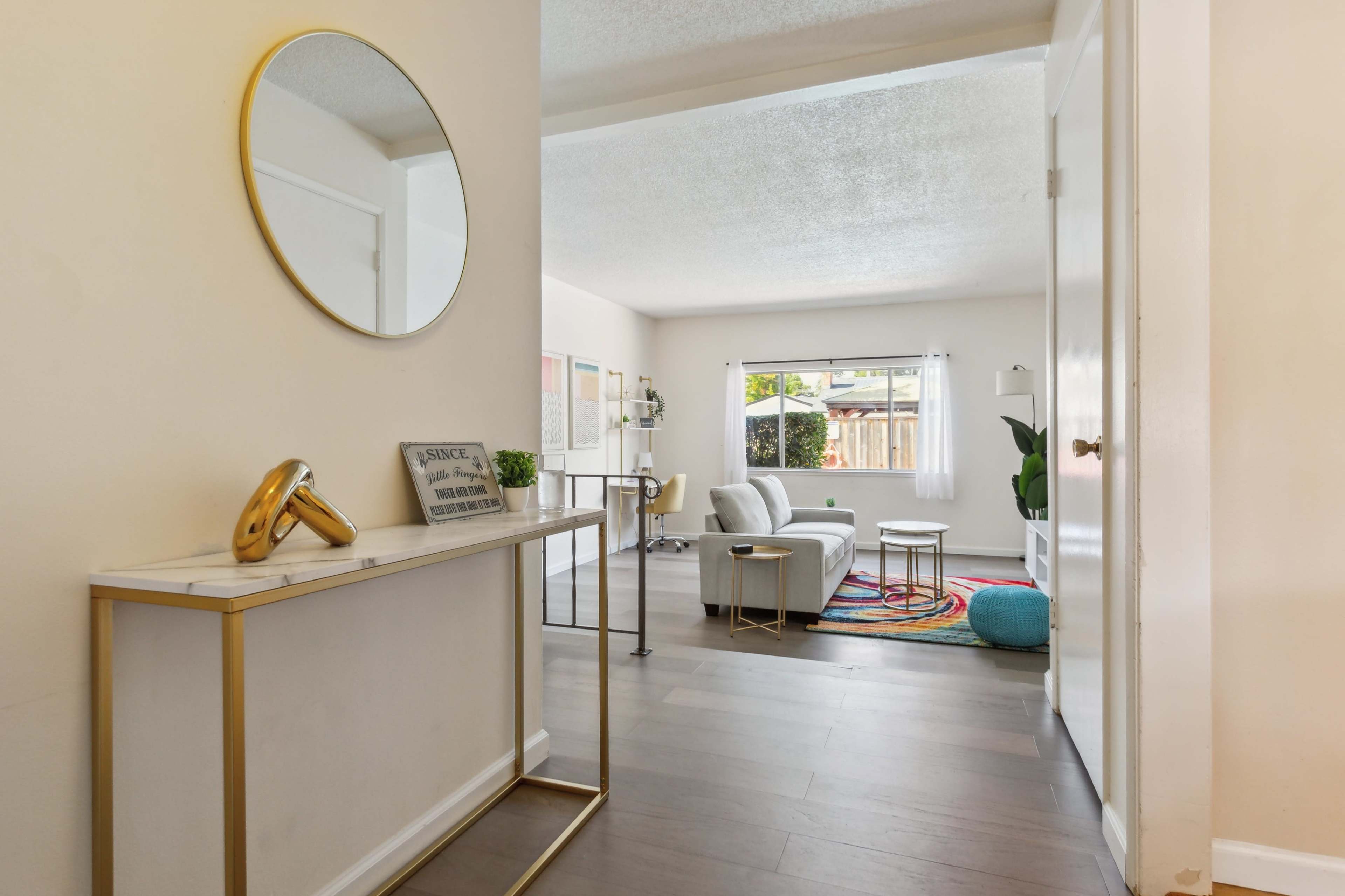 A bright hallway leading into a living room, featuring a console table with a round mirror and a comfortable seating area with a colorful rug.