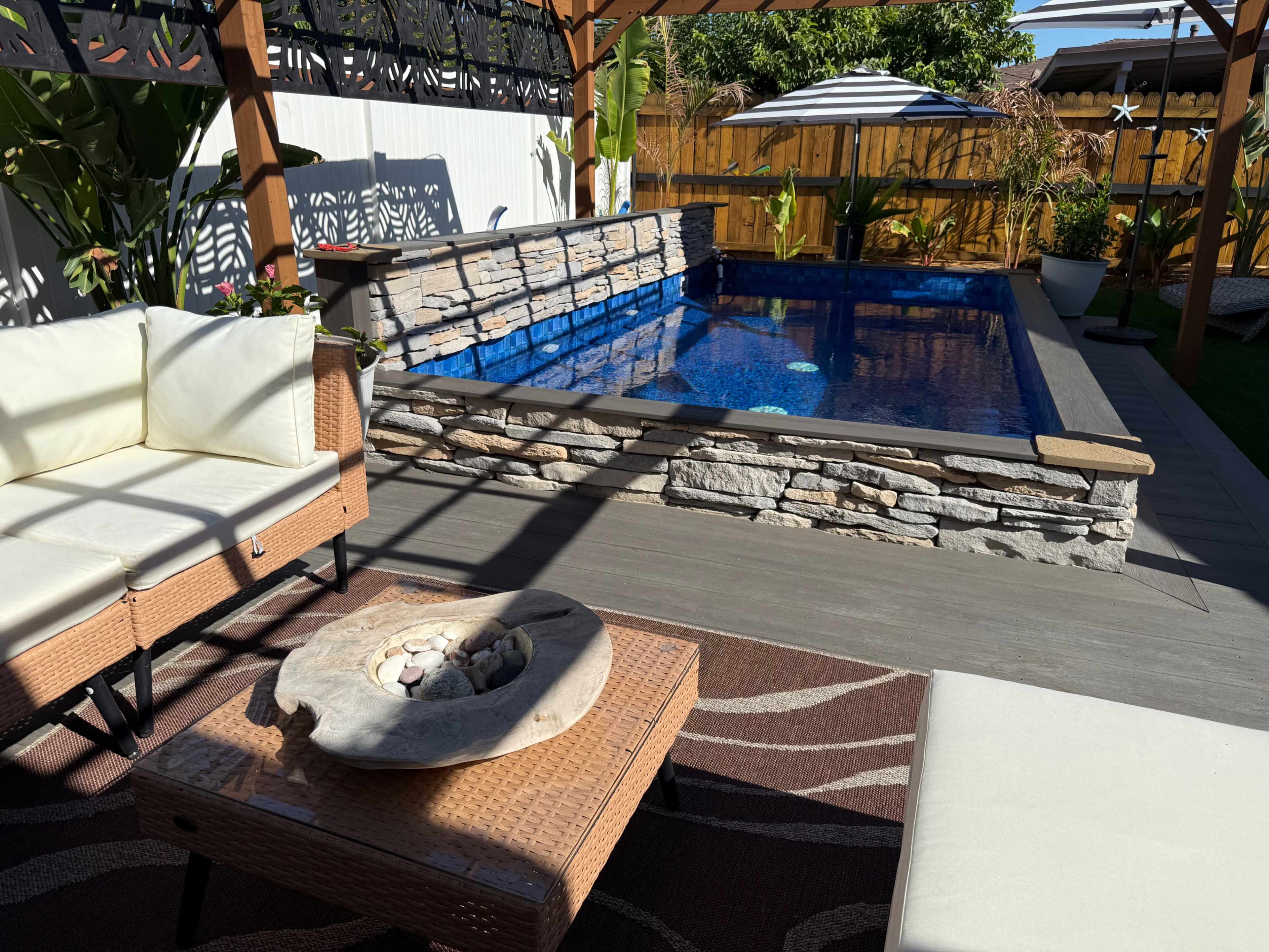 The image depicts a backyard pool area featuring a stone-accented pool, a shaded seating area with two white chairs, and decorative landscaping.