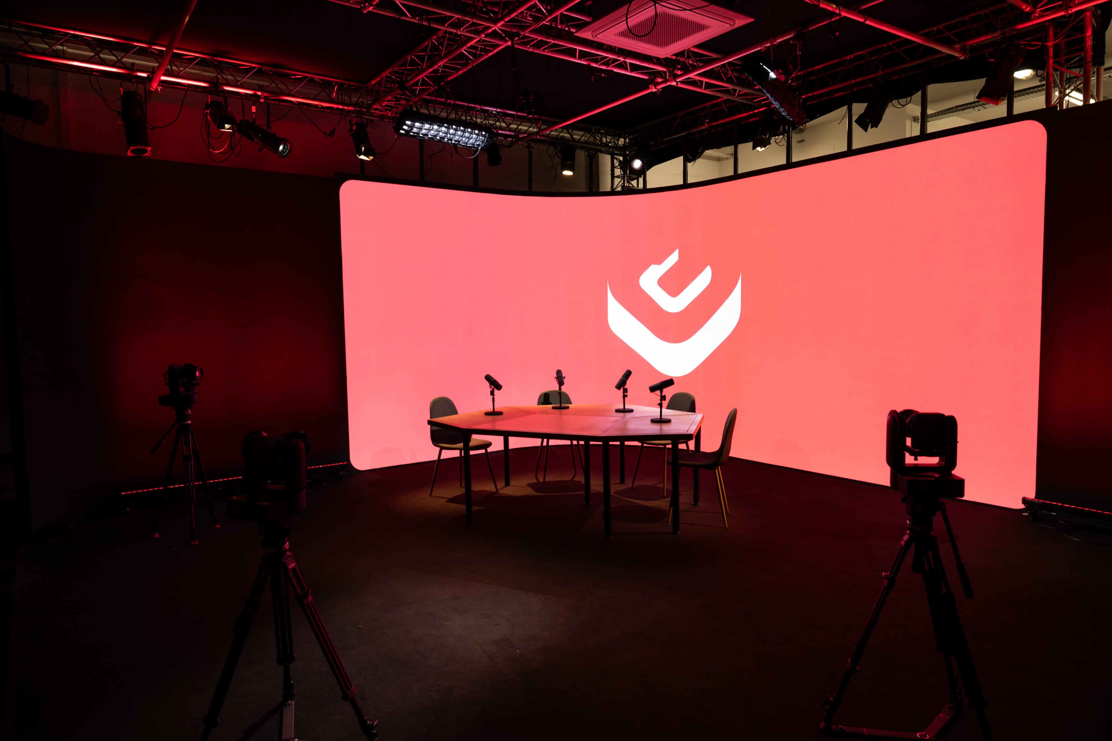 A modern studio setup features a round table with microphones and cameras in front of a large red backdrop displaying a logo.