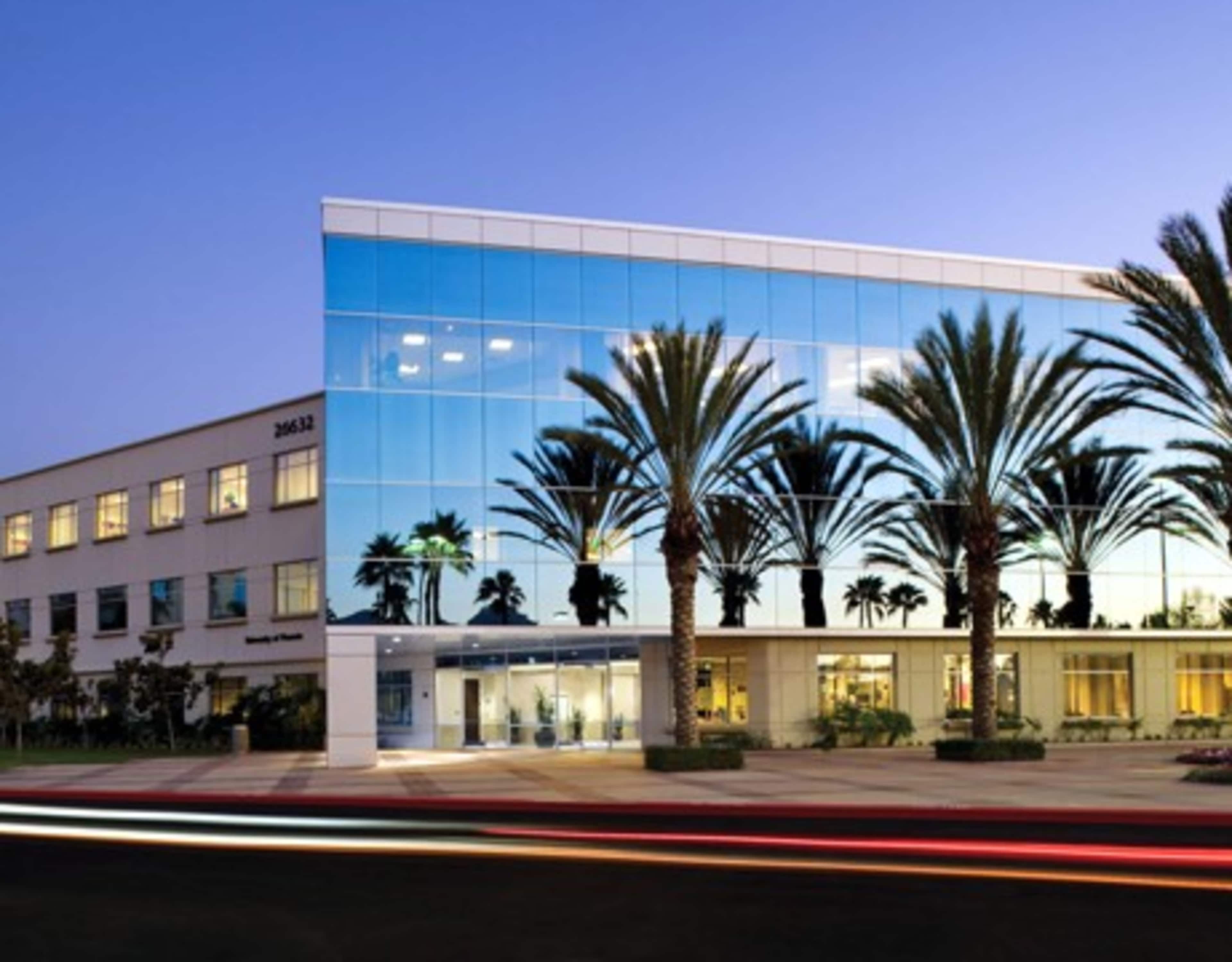 The image shows a modern, multi-story building with large glass windows reflecting palm trees and a street in the foreground at dusk.