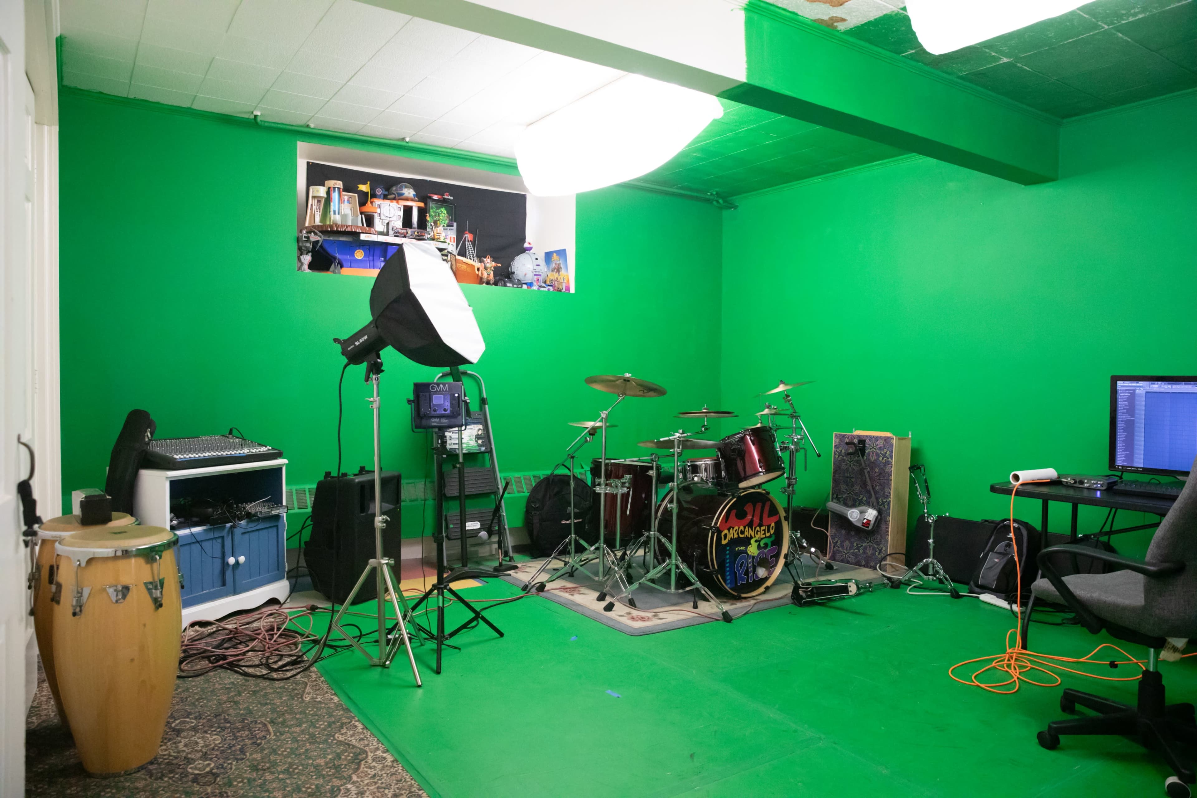The image shows a green-painted room set up as a music studio, featuring a drum set, conga drums, recording equipment, and a computer.