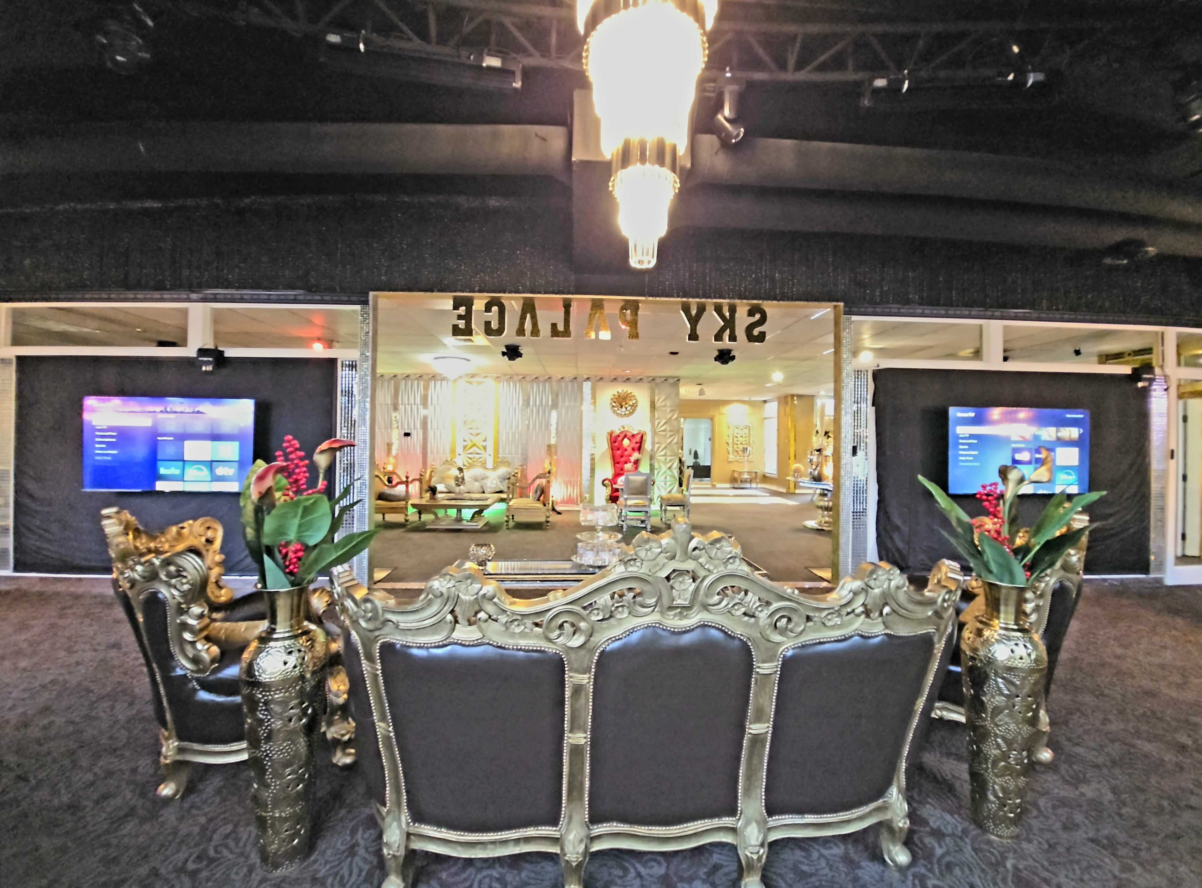 The image shows a lavish interior with ornate gold furniture facing a large mirror, flanked by two screens displaying colorful visuals.