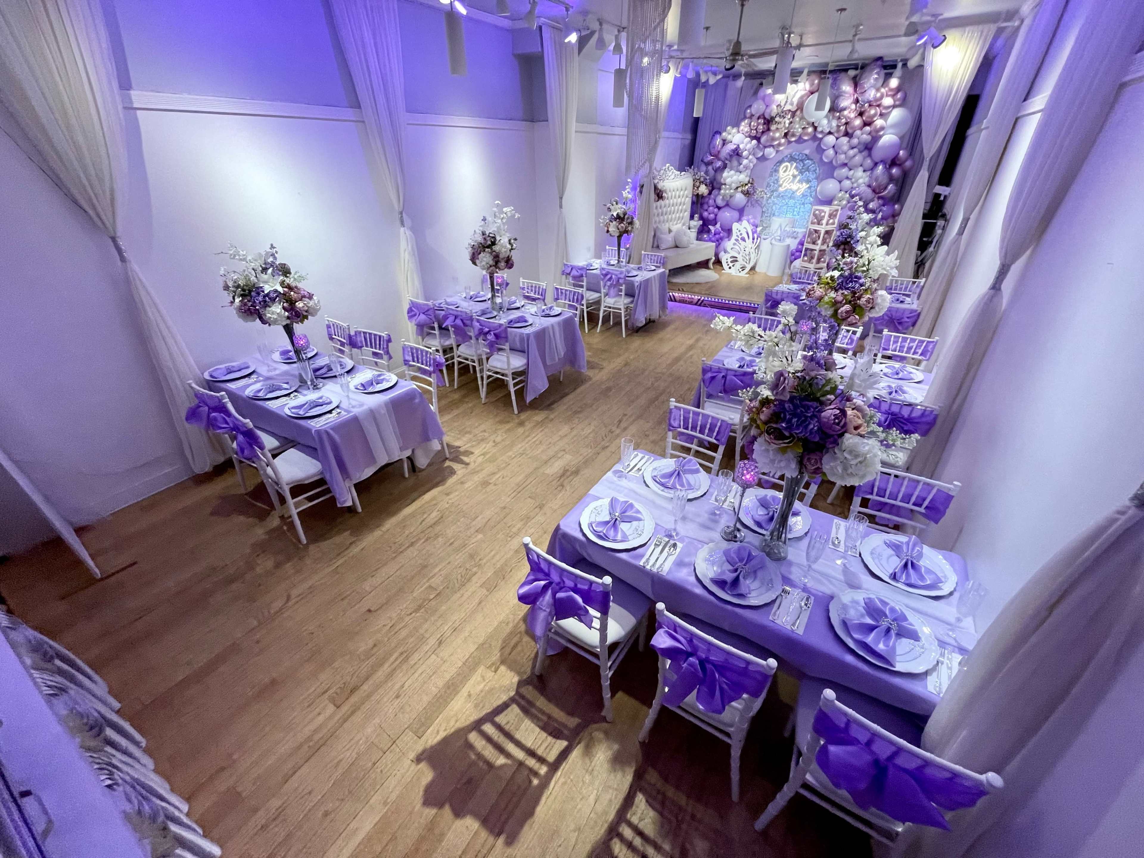 The image shows a banquet hall decorated with purple-themed table settings and floral centerpieces, featuring multiple tables arranged for an event.