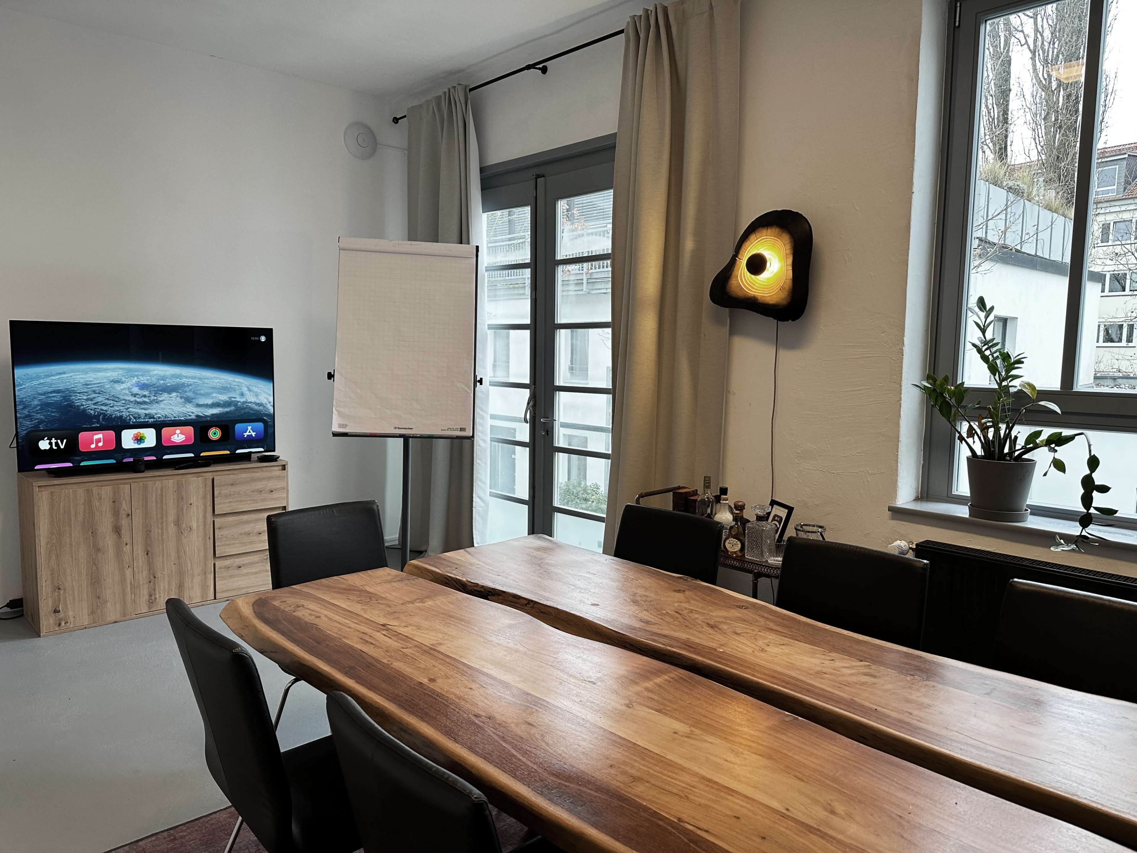 A spacious meeting room features a large wooden table, a TV mounted on the wall, a flip chart stand, and a potted plant near a window.