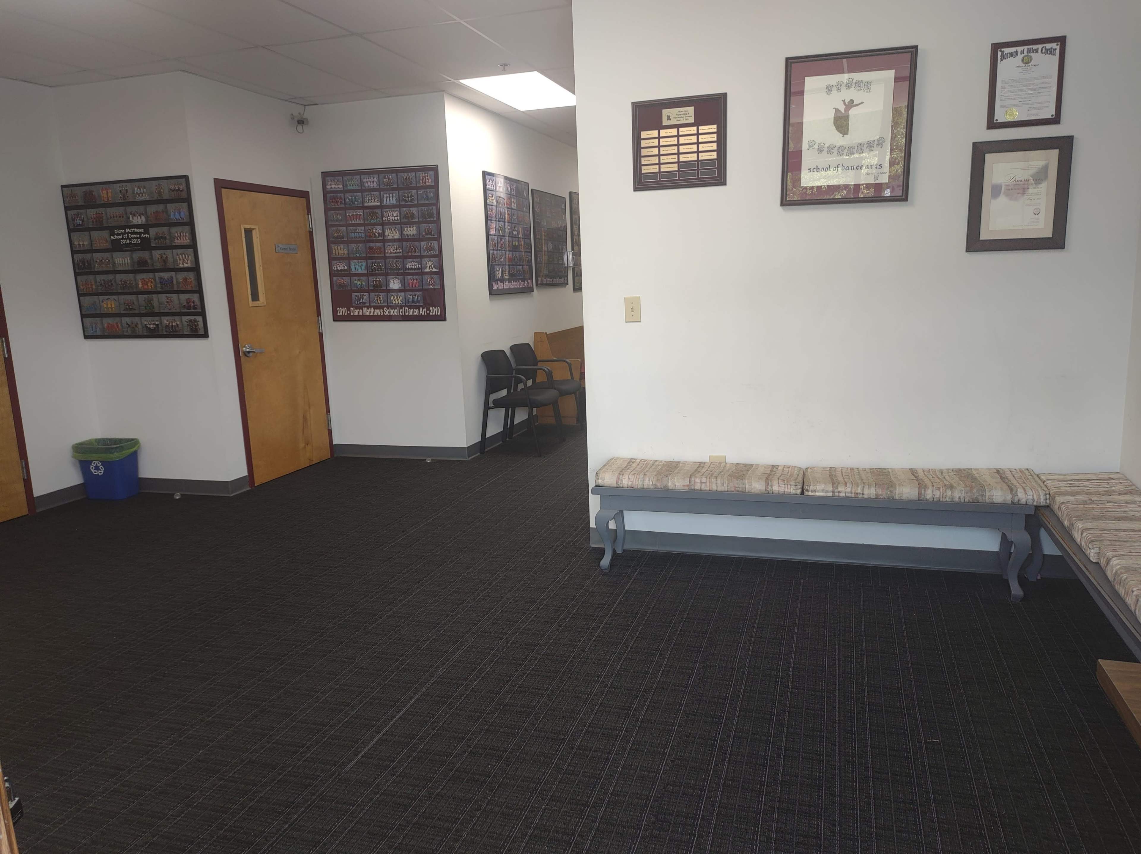 The image shows a waiting area with a wall-mounted display of plaques and framed photographs, a long bench against one wall, and a few chairs arranged nearby.