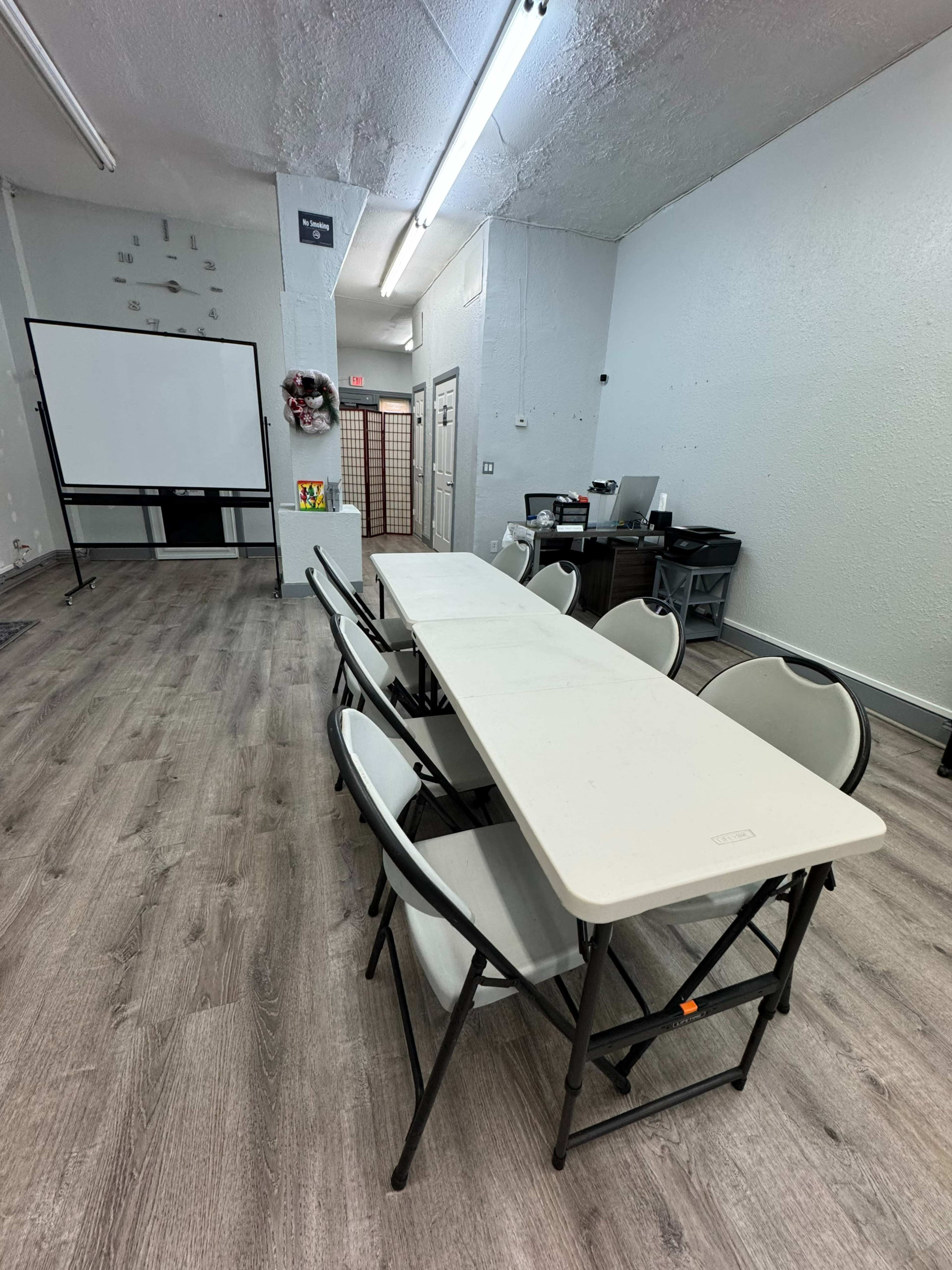 A room with light-colored walls contains a long table with foldable chairs, a projector screen, and a workspace area in the background.
