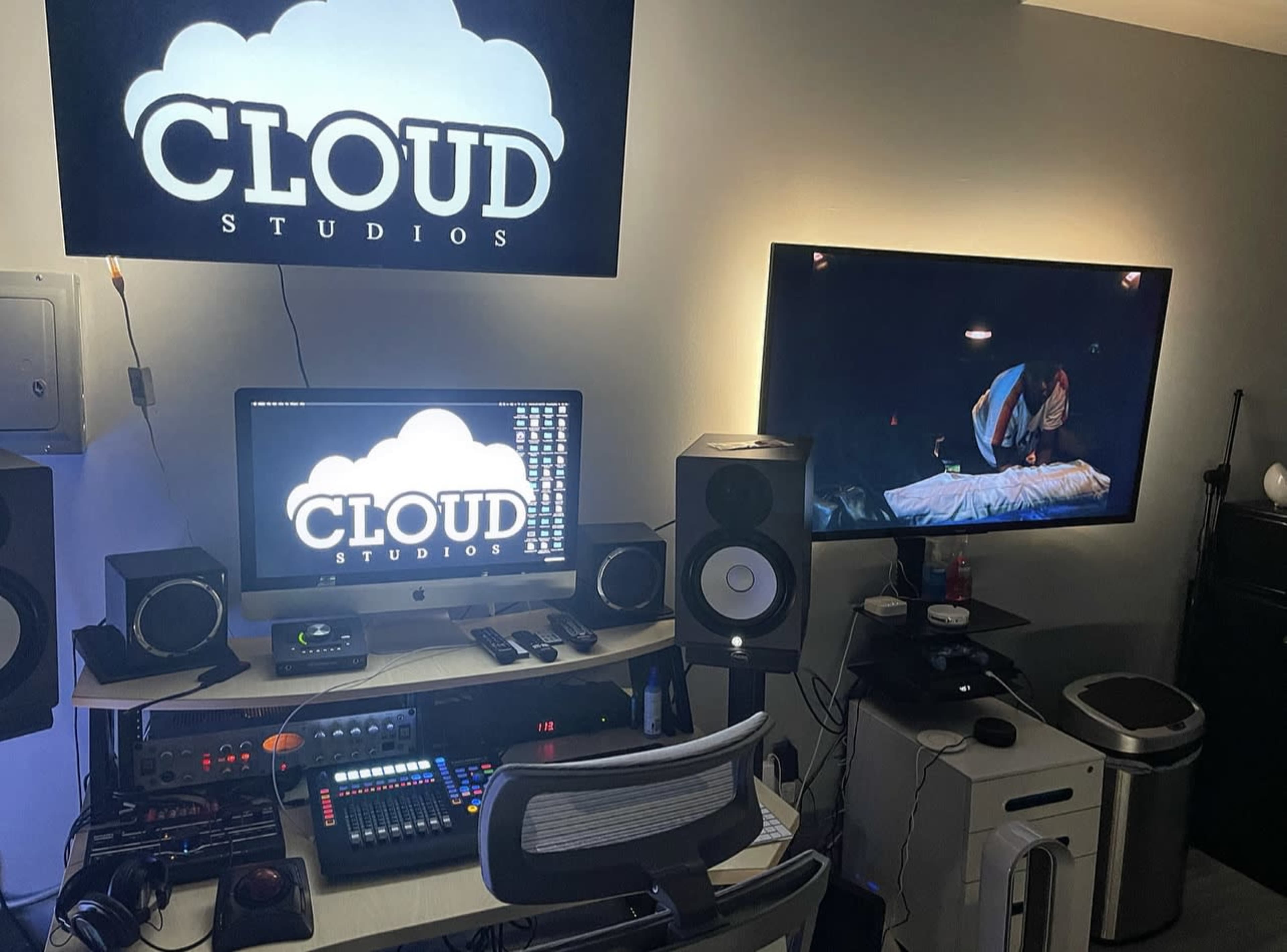 A music studio setup featuring a computer, audio equipment, and large screens displaying a logo and a scene.