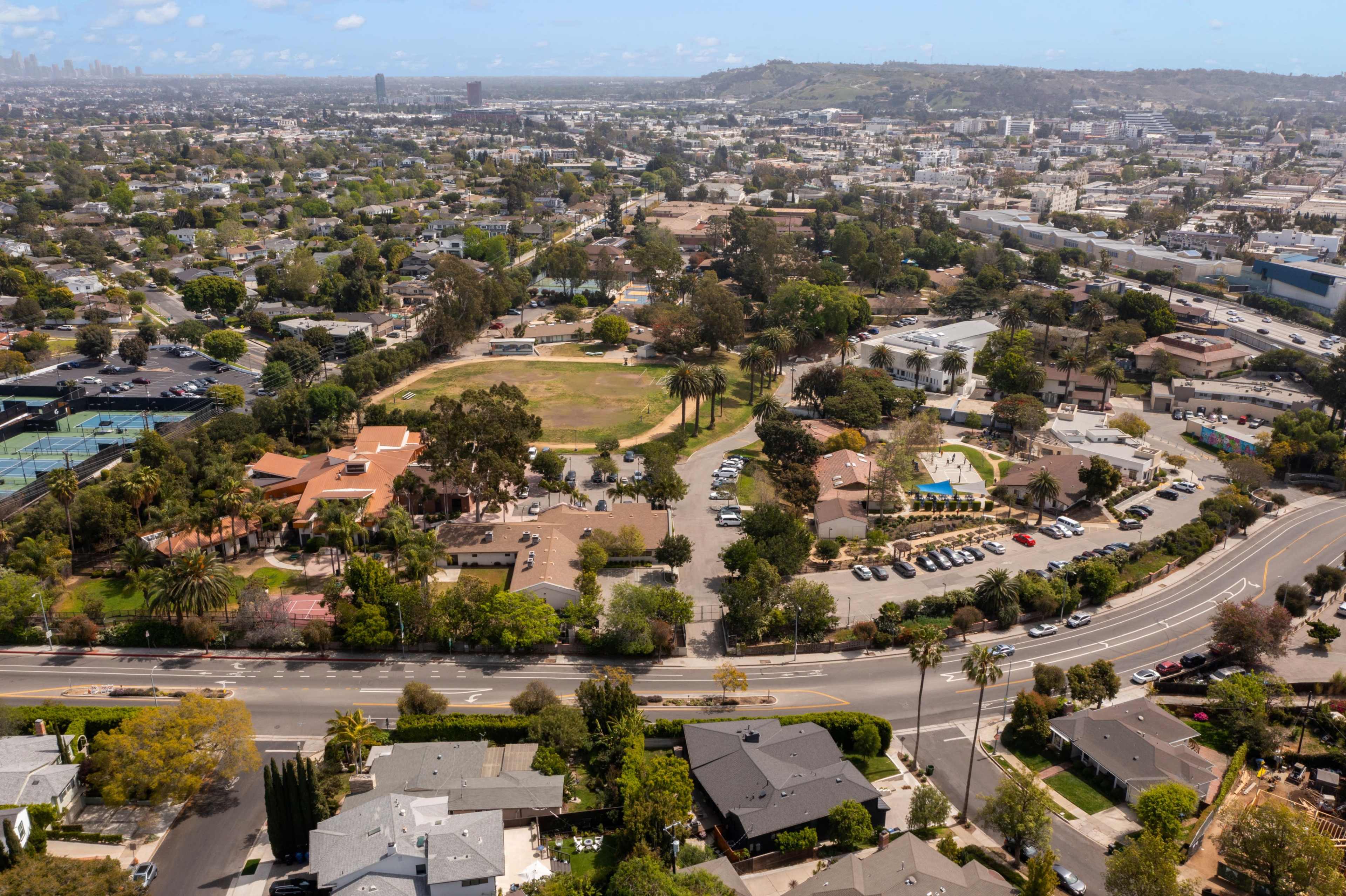 An aerial view of a suburban area with a park, houses, and a winding road, surrounded by greenery and urban structures.