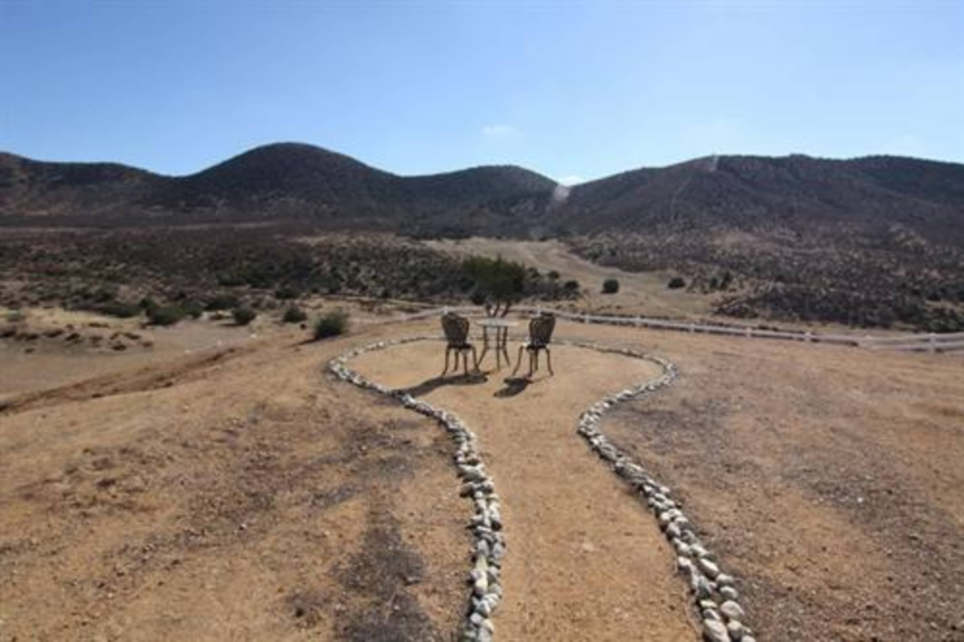 Two chairs and a small table are situated on a gravel path that winds through a dry, mountainous landscape.