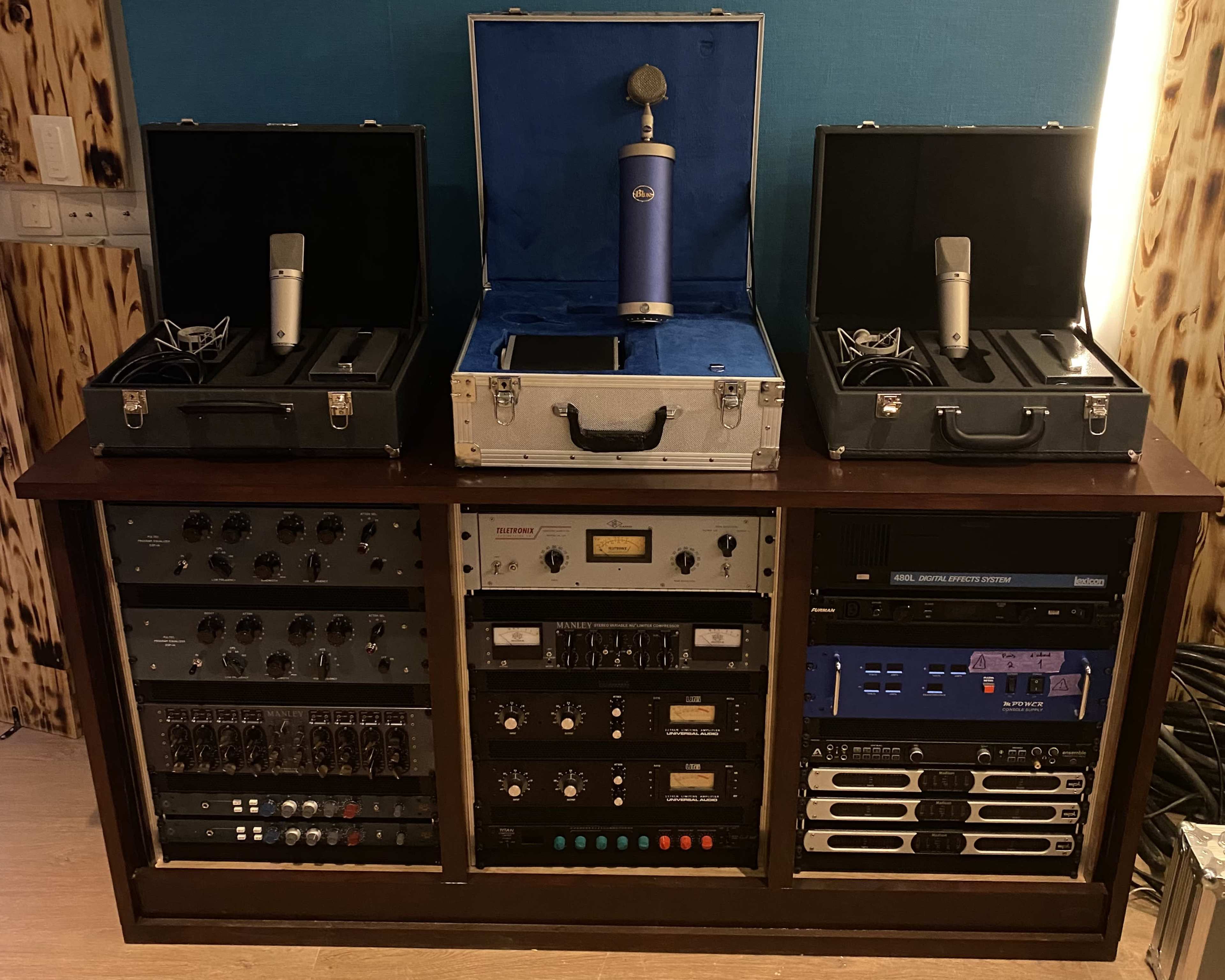 A wooden audio equipment rack containing various microphones, interfaces, and signal processors neatly organized with two microphone cases on top.
