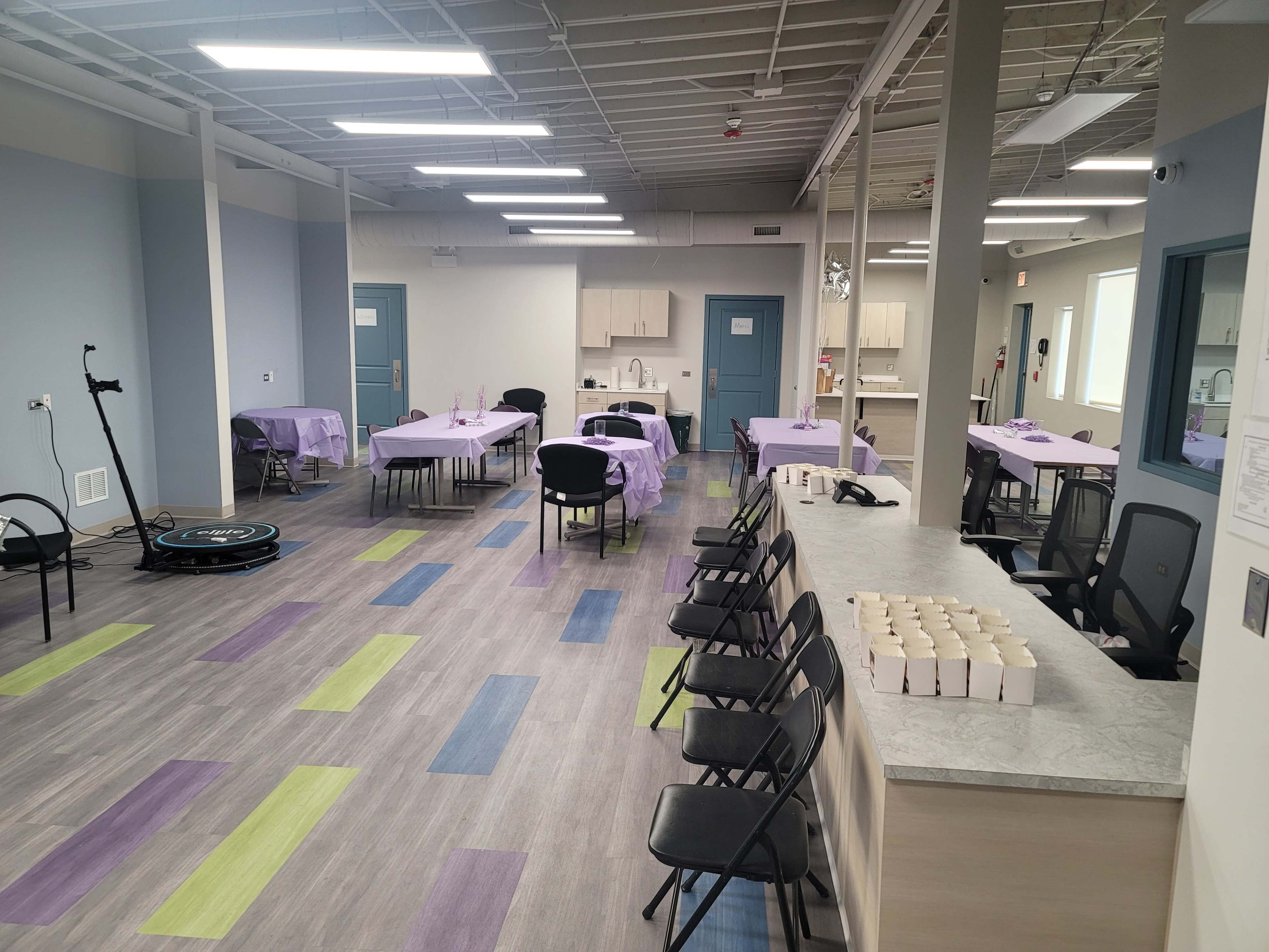 A spacious room with multiple tables covered in purple tablecloths, chairs arranged around them, and a kitchenette area visible in the background.