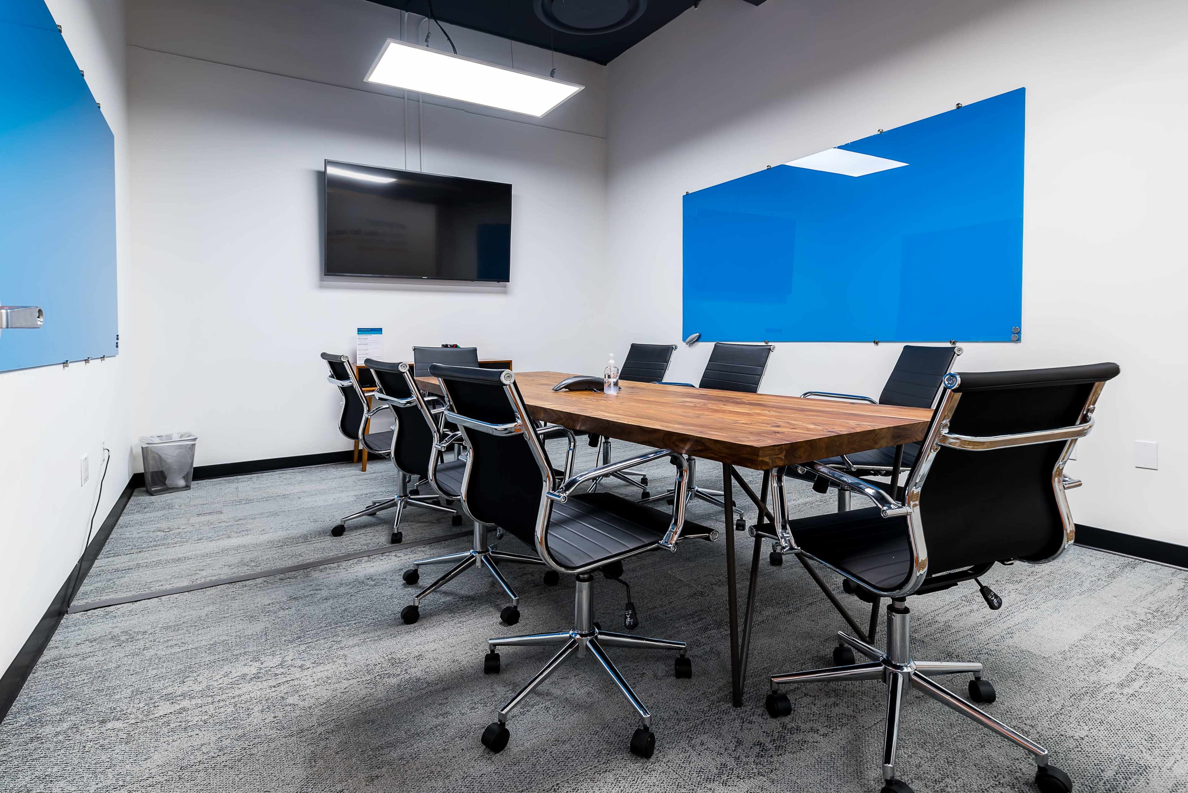 The image shows a modern conference room featuring a wooden table surrounded by black chairs, with a television on the wall and blue whiteboards.