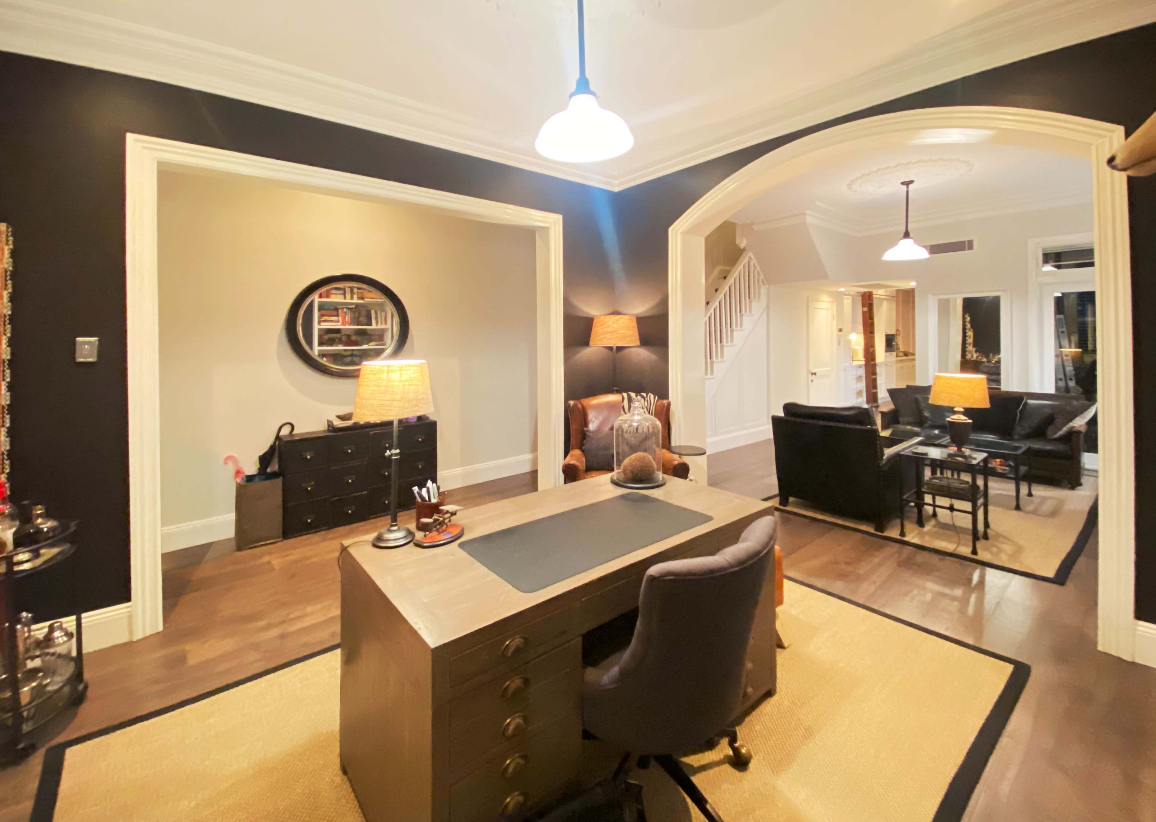 The image shows an elegantly designed office space featuring a wooden desk, two armchairs, and a staircase leading to another area.