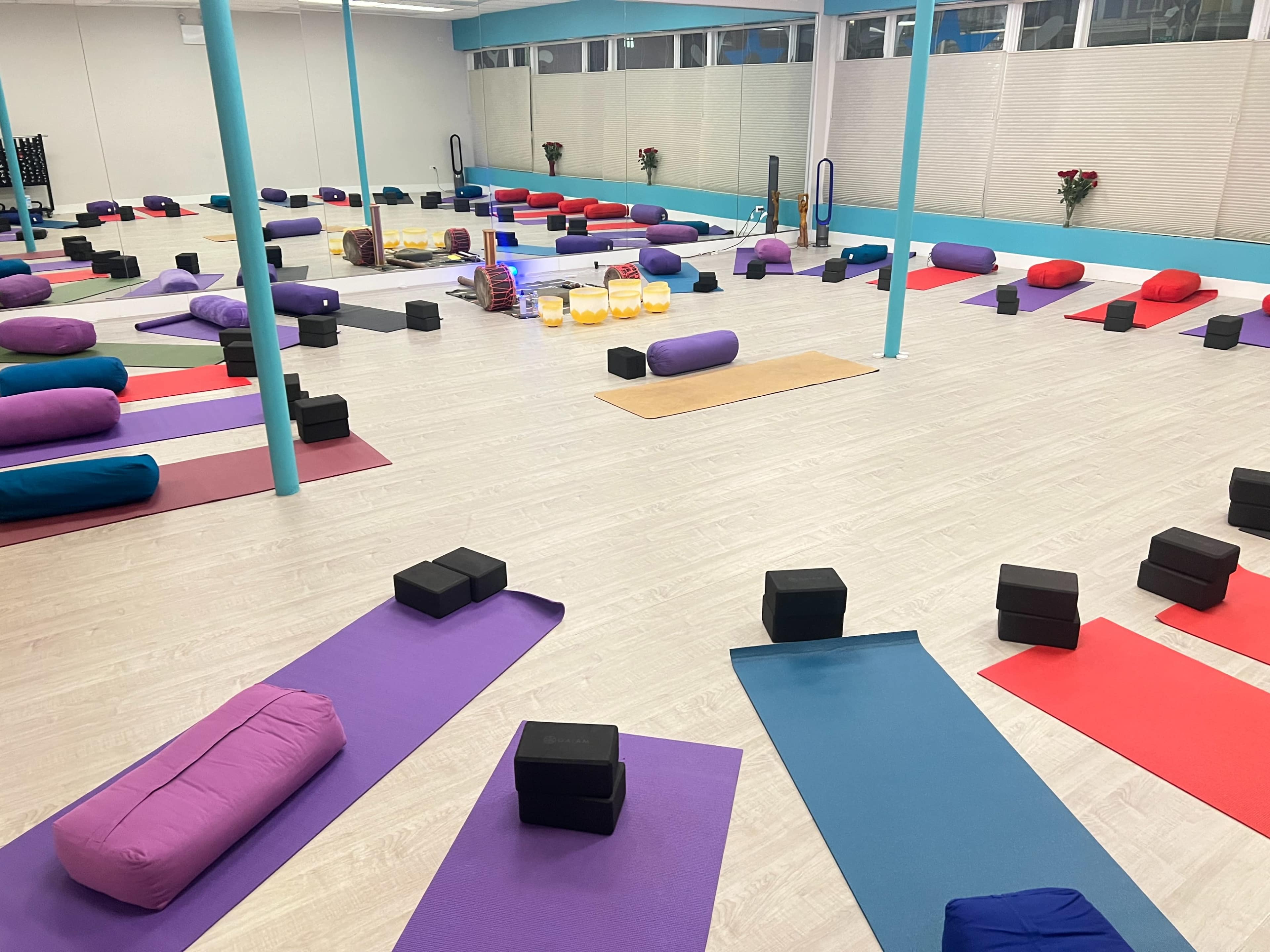 The image shows a spacious yoga studio with various colored mats, bolsters, and blocks arranged in a symmetrical layout.