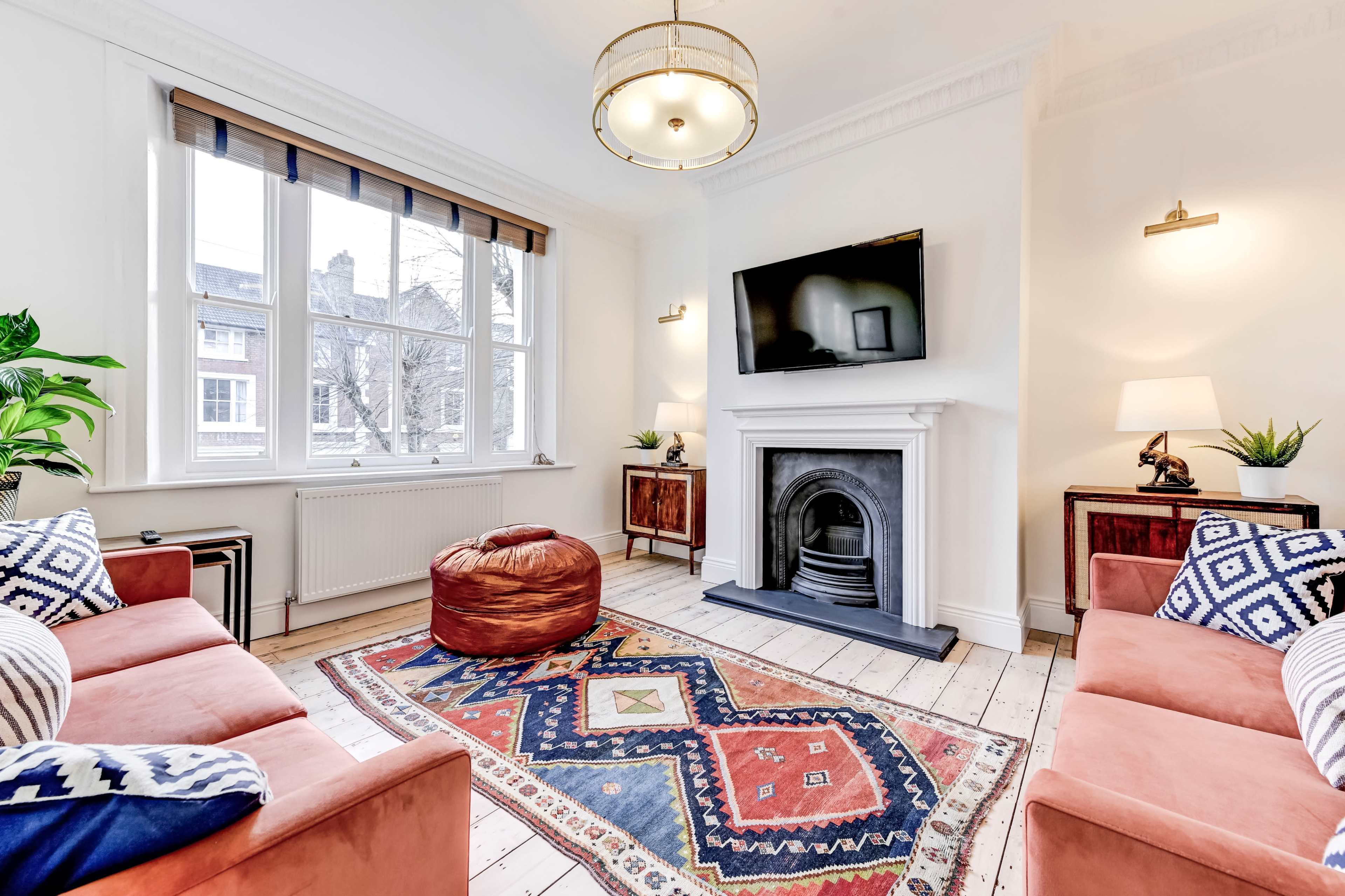 The living room features two orange sofas facing a central rug, a television mounted above a white fireplace, and large windows allowing natural light to enter.