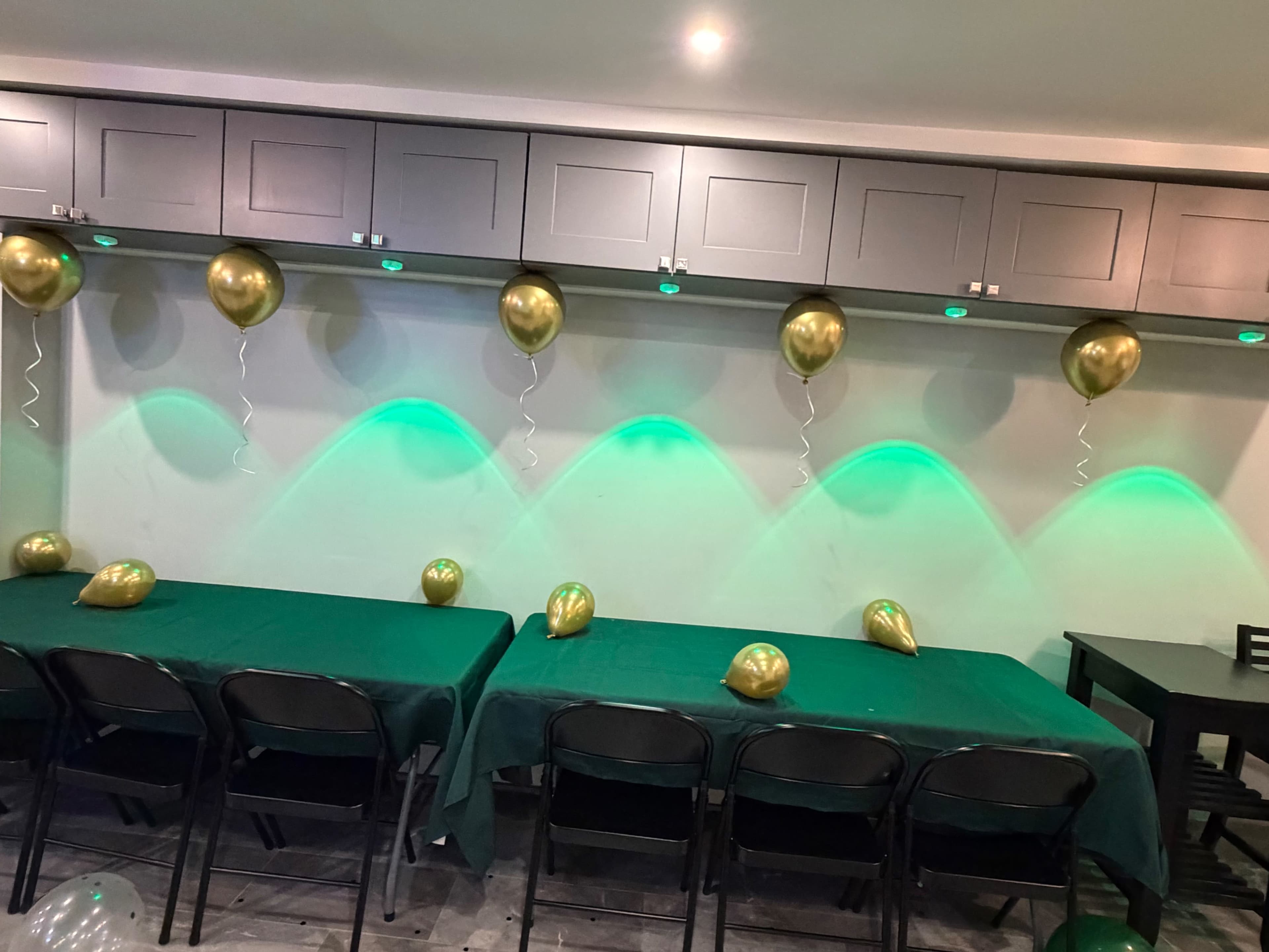 The scene features a decorated indoor space with green tablecloths, black chairs, and gold balloons arranged along a wall illuminated by soft green lighting.