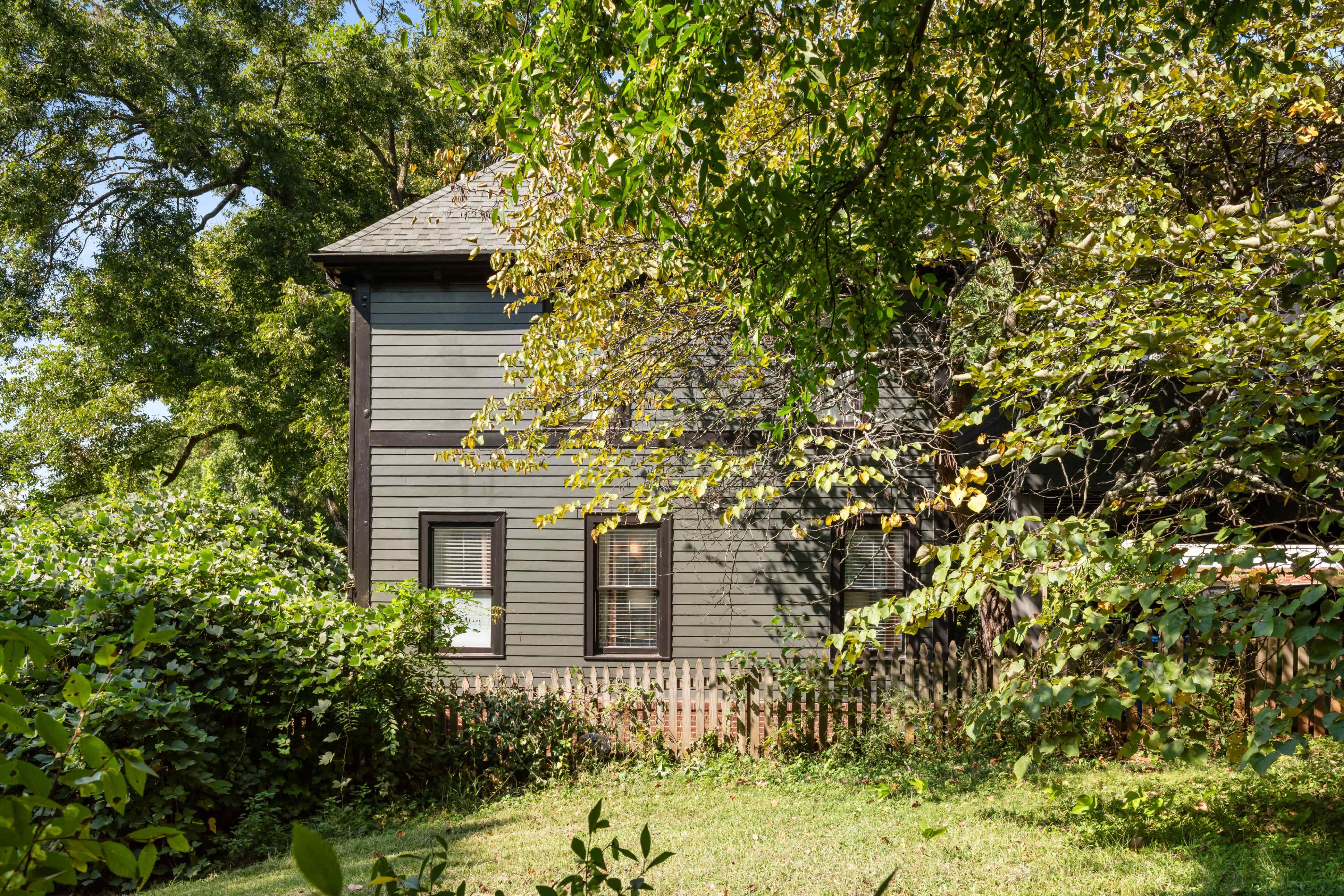 A two-story house with gray siding is partially obscured by green foliage and trees in a residential yard.