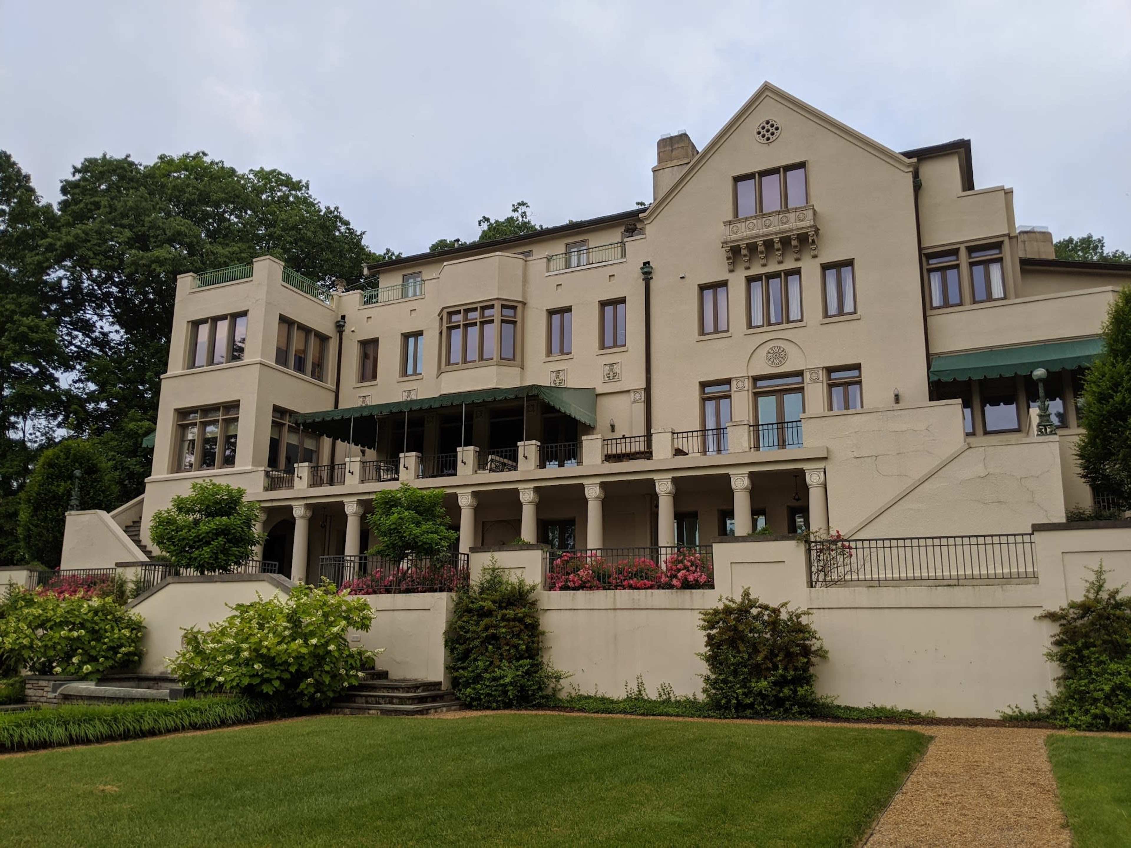 The image shows a large, multi-level mansion with a terrace overlooking a landscaped garden, featuring decorative architecture.