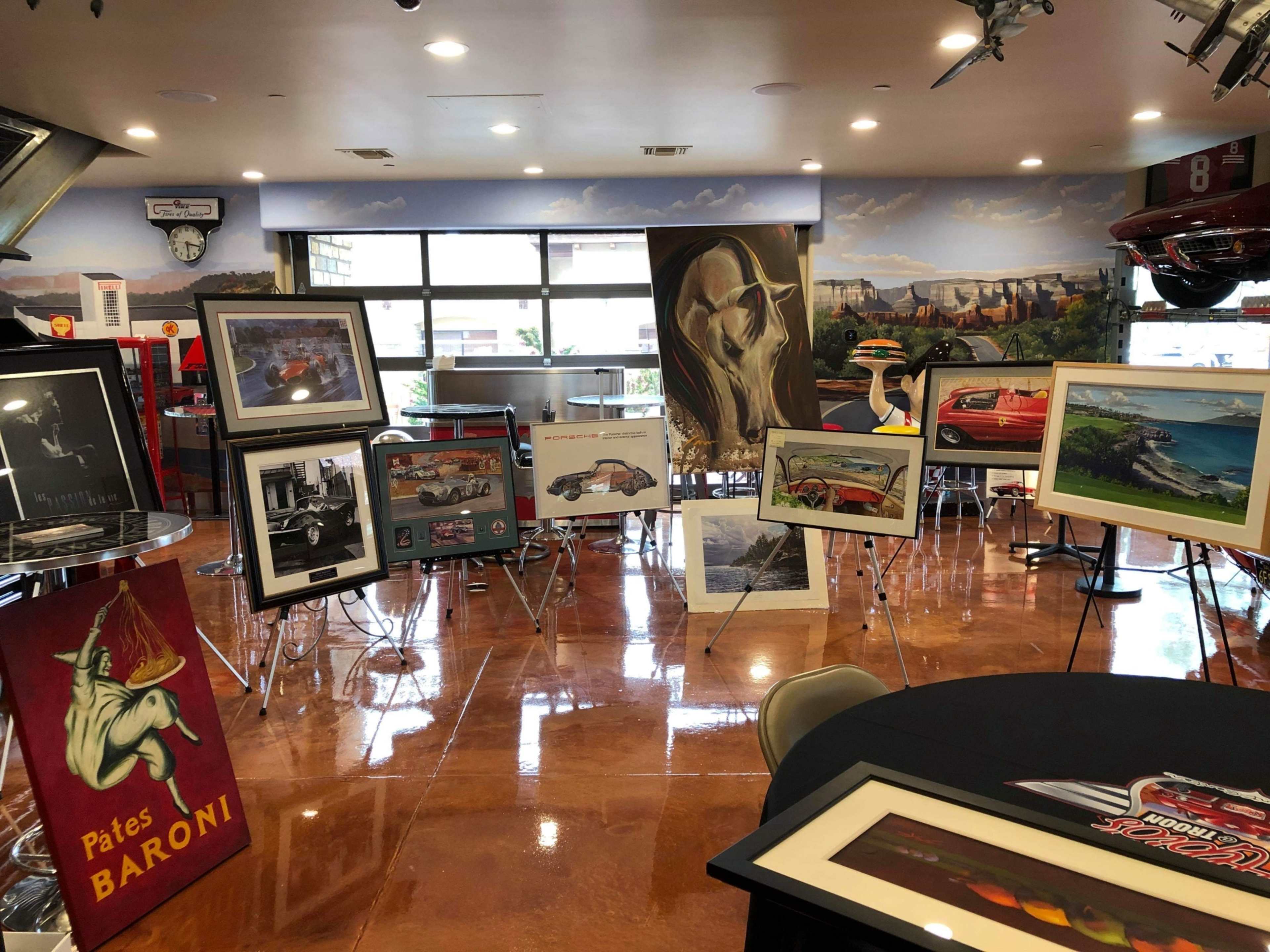 An interior space with various framed artworks displayed on easels, alongside a round table and vintage car memorabilia.