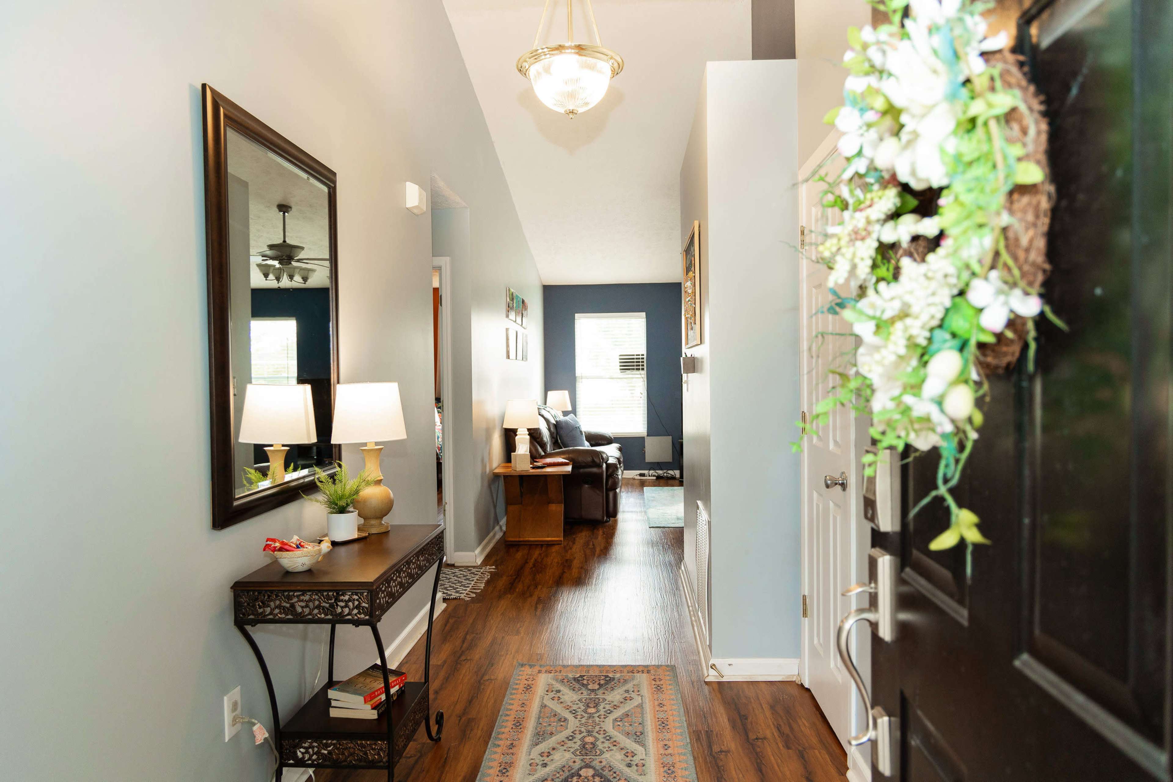A hallway features a console table with a lamp and decorative items, leading toward a living area with a sofa and large window.