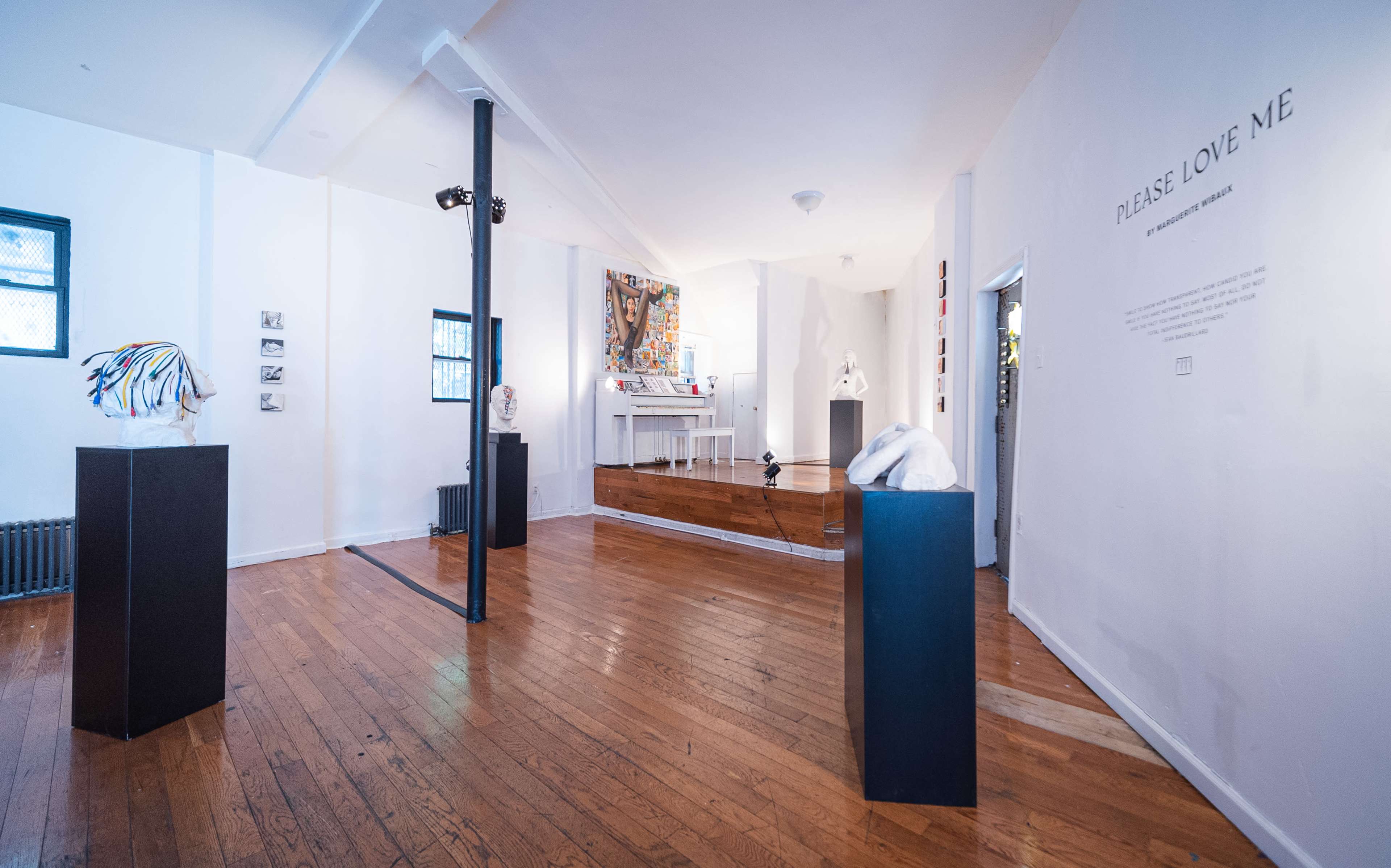 An art gallery featuring sculptures displayed on pedestals in a well-lit room with wooden flooring and a text installation on the wall.