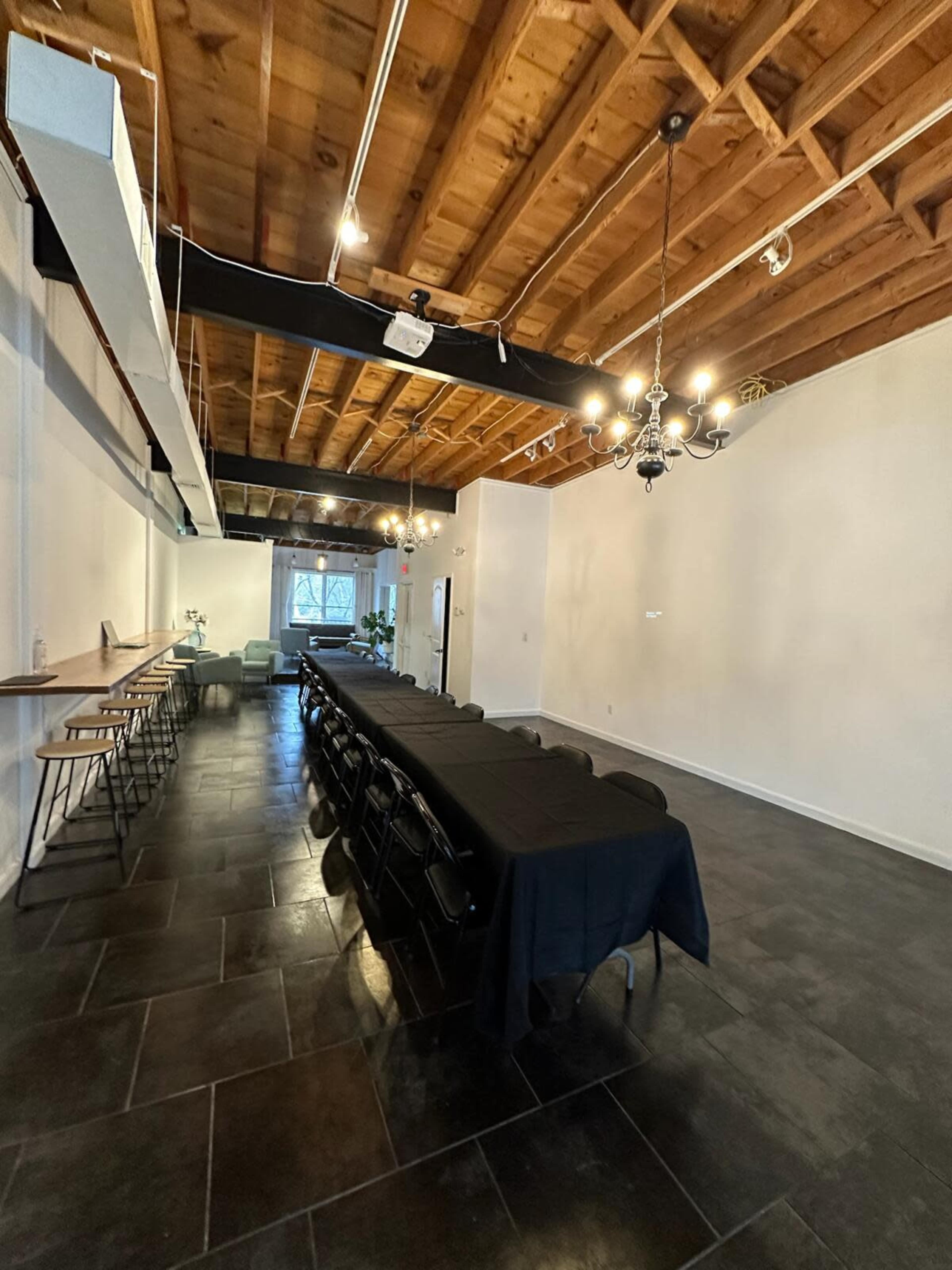A spacious, well-lit event room featuring a long black table with chairs, a bar area along one wall, and a chandelier hanging from the ceiling.