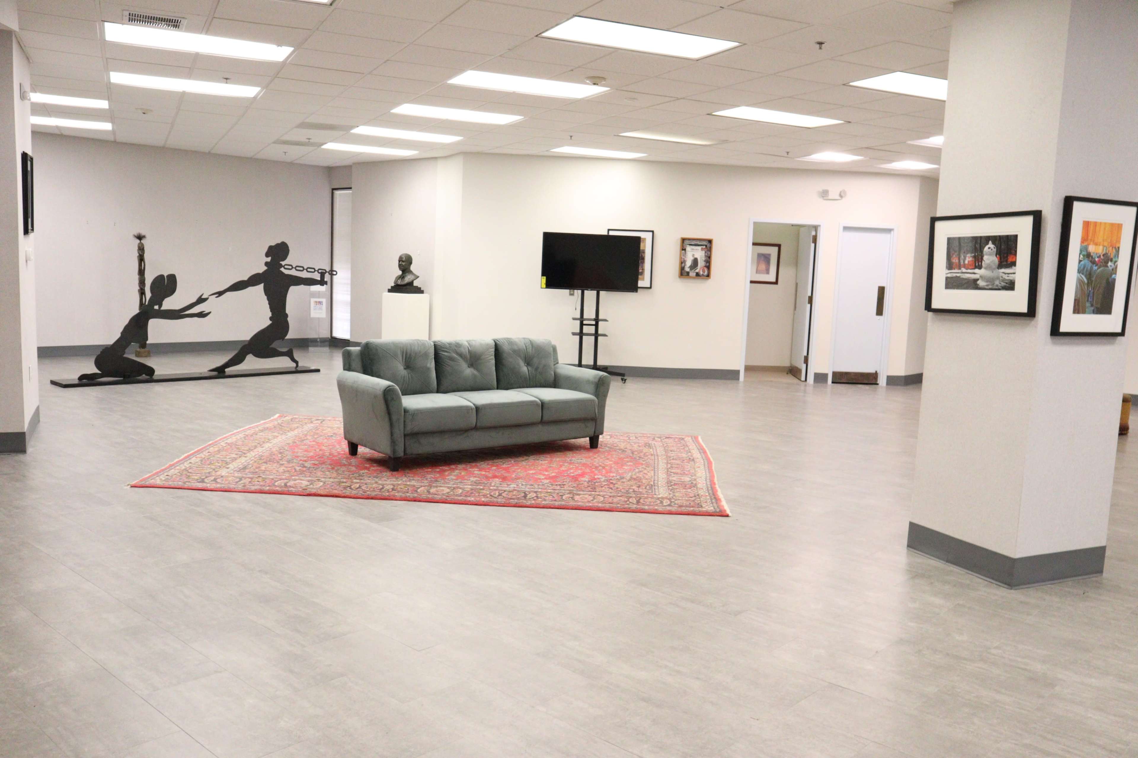 A spacious room with a gray couch on a red area rug, accompanied by artwork on the walls and a large black silhouette sculpture in the corner.